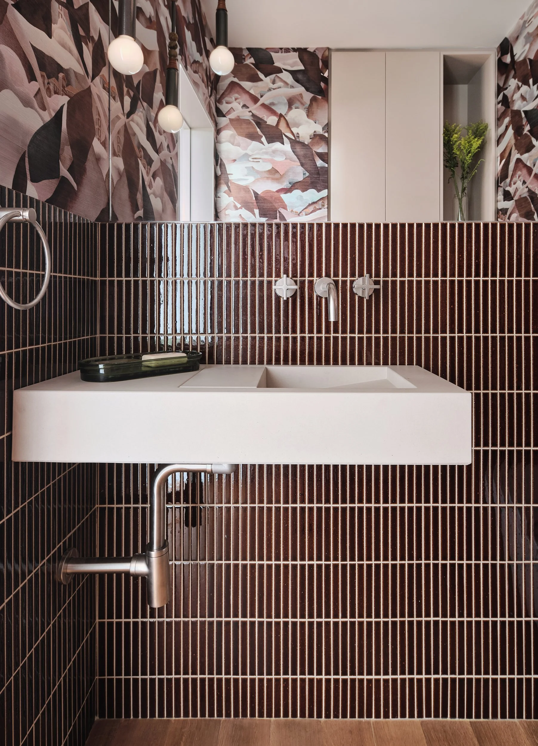 A modern bathroom with dark brown vertical tile walls, a white rectangular sink, and a bronze finish faucet. There is a black soap dish on the sink. The background has patterned wallpaper with abstract leaf and mountain designs and a small cabinet with a green plant. Hanging light bulbs are on the ceiling.