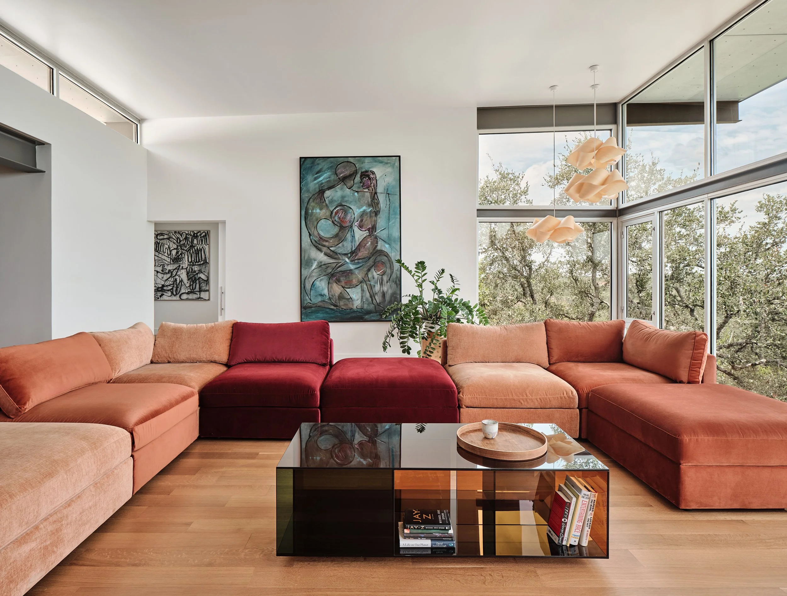 Modern living room with large L-shaped sectional sofa in shades of pink and red, mid-century style black glass coffee table with books and a wooden tray, large window with a view of trees outside, white walls with artwork, potted plant, and contemporary pendant lights.