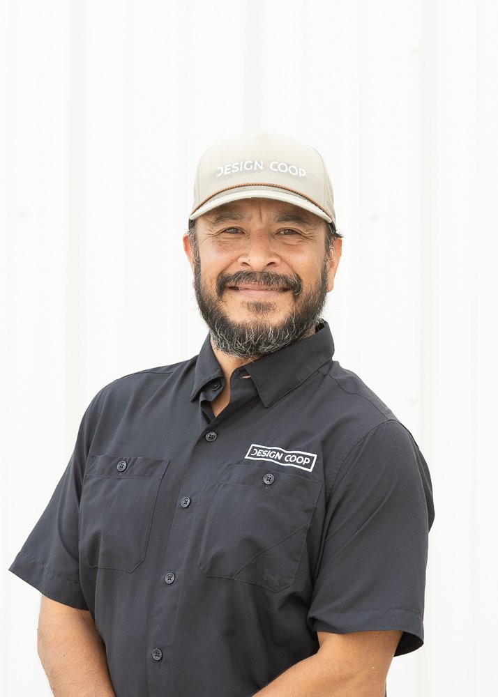 A man with short dark hair smiling, wearing a black polo shirt with a small logo that says 'Design Coop' on the left chest, standing against a plain white background.