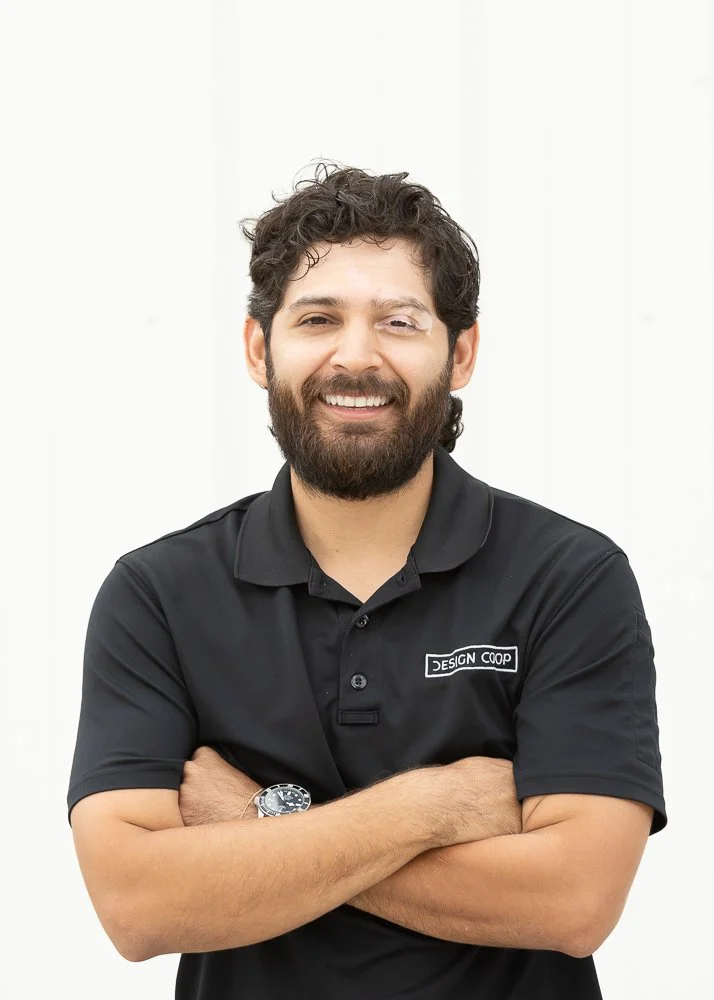 Man with dark hair and beard wearing a black polo shirt with a 'Design Coop' logo on the chest, smiling, standing against a white background.