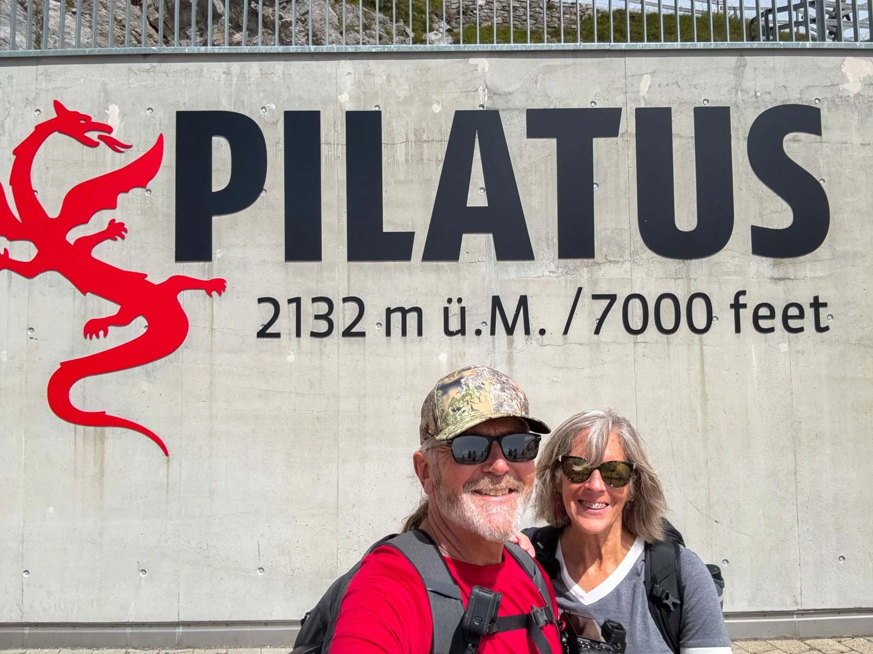 Two smiling hikers, a man and a woman, standing in front of a large sign for Pilaus mountain, with the elevation of 2132 meters or 7000 feet, and a red dragon logo. The man is wearing sunglasses, a camo hat, a red shirt, and a backpack. The woman is 