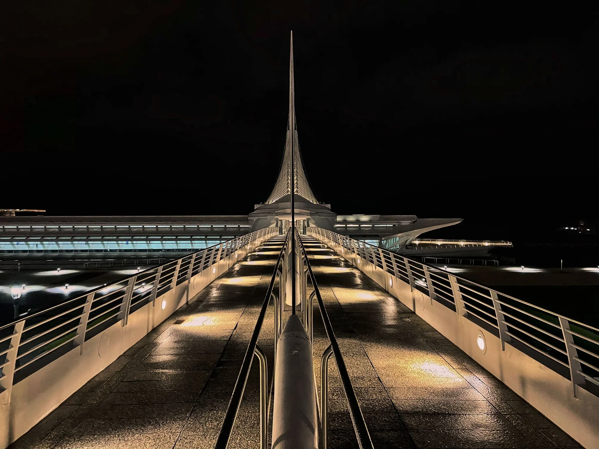 Night view of a modern architectural building with a pointed spire and a walkway with railing leading to it.