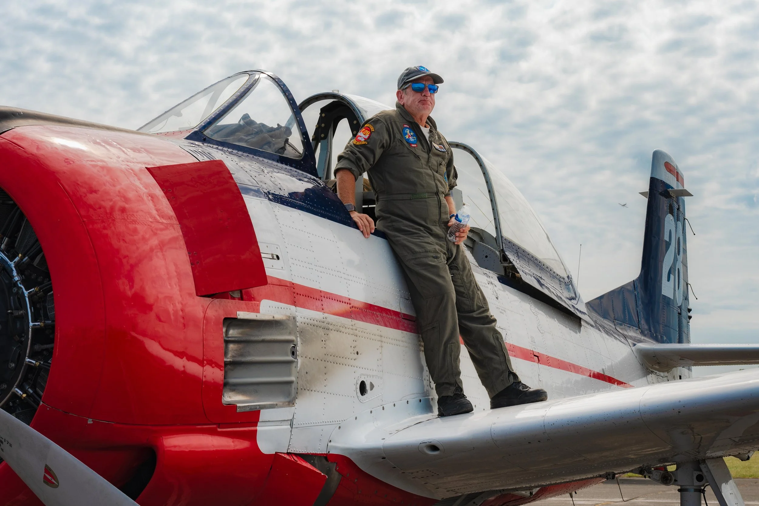 A man in a flight suit with patches and sunglasses is standing on an airplane's wing, holding a water bottle, with a cloudy sky in the background.