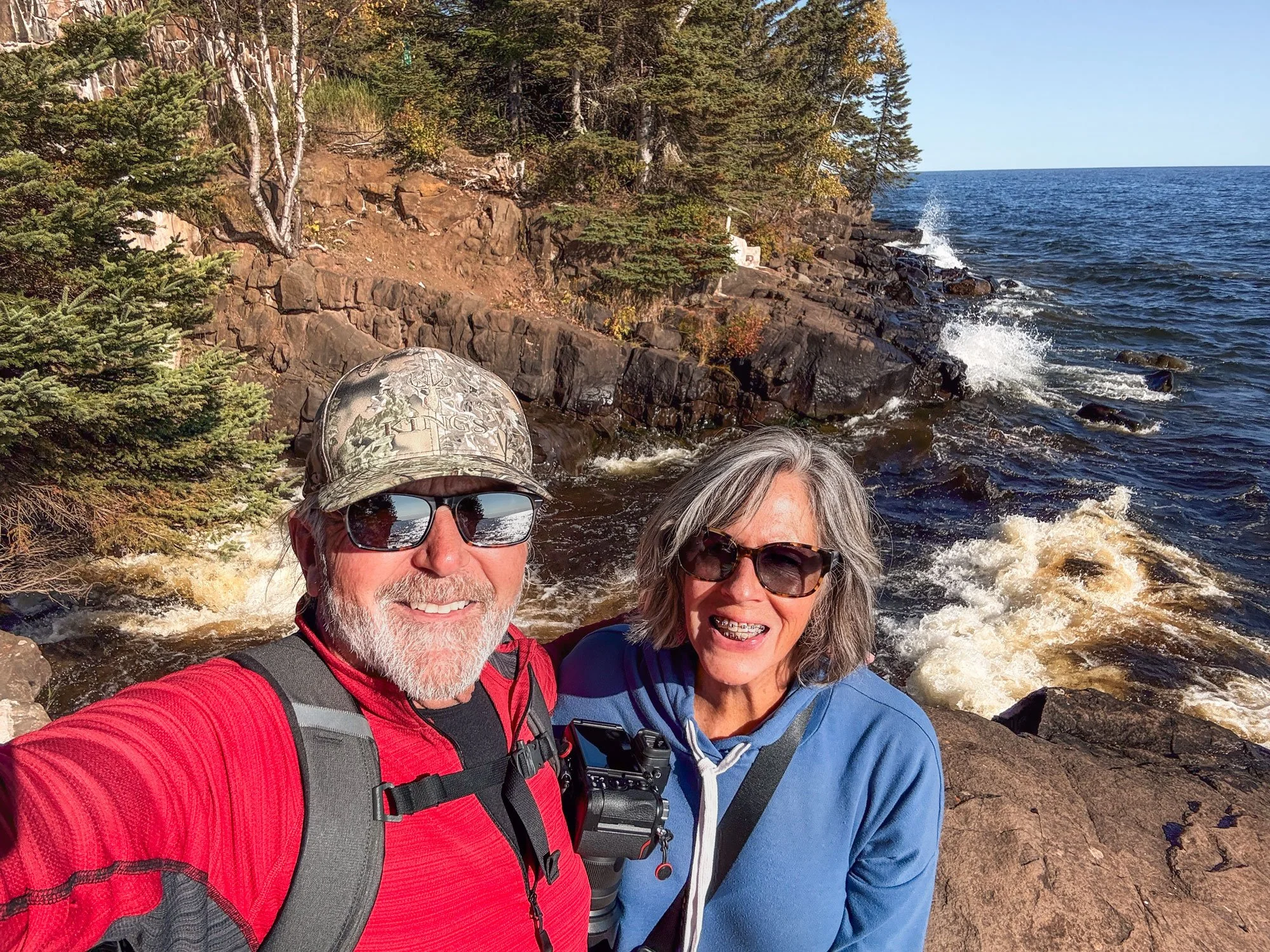 A smiling middle-aged man and woman wearing sunglasses and outdoor clothing taking a selfie by a rocky shoreline with waves and trees in the background.