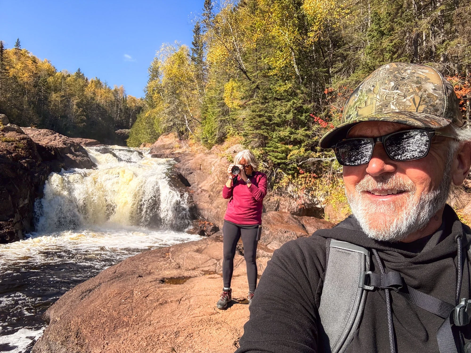 Older man with a beard wearing sunglasses, a camo hat, and a backpack taking a selfie outdoors near a waterfall. Woman in a red jacket with sunglasses and camera standing on rocks nearby in a forested area during fall.