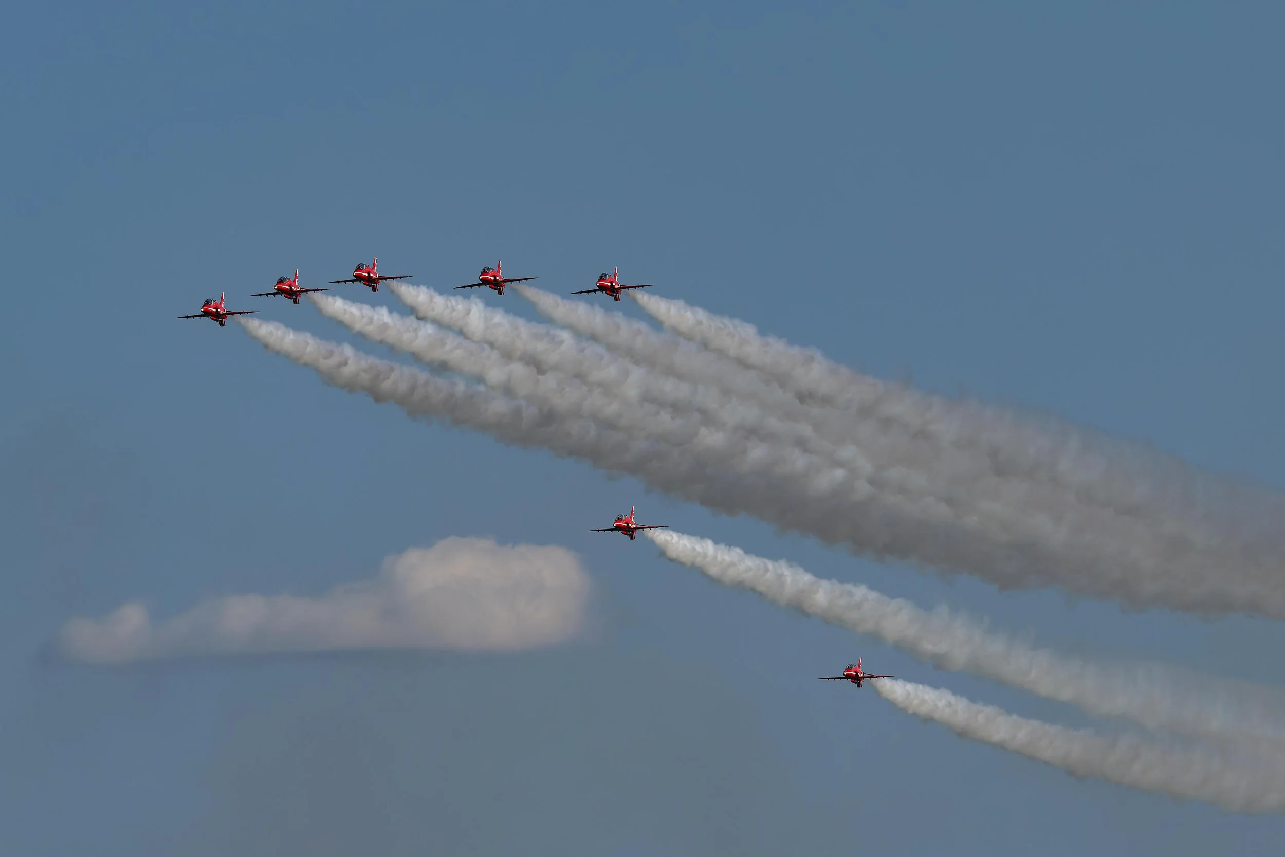 Five red aircraft flying in a formation, trailing white smoke against a blue sky.
