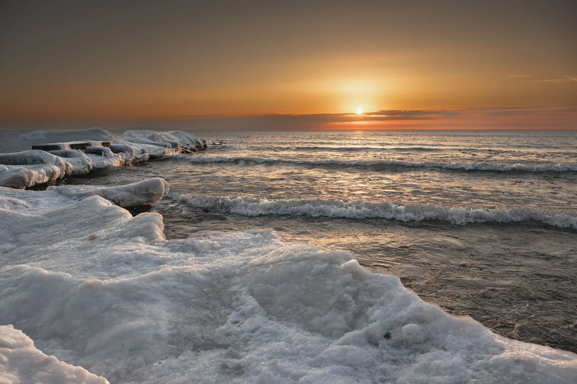 Sunset over a frozen ocean with ice-covered rocks in the foreground.