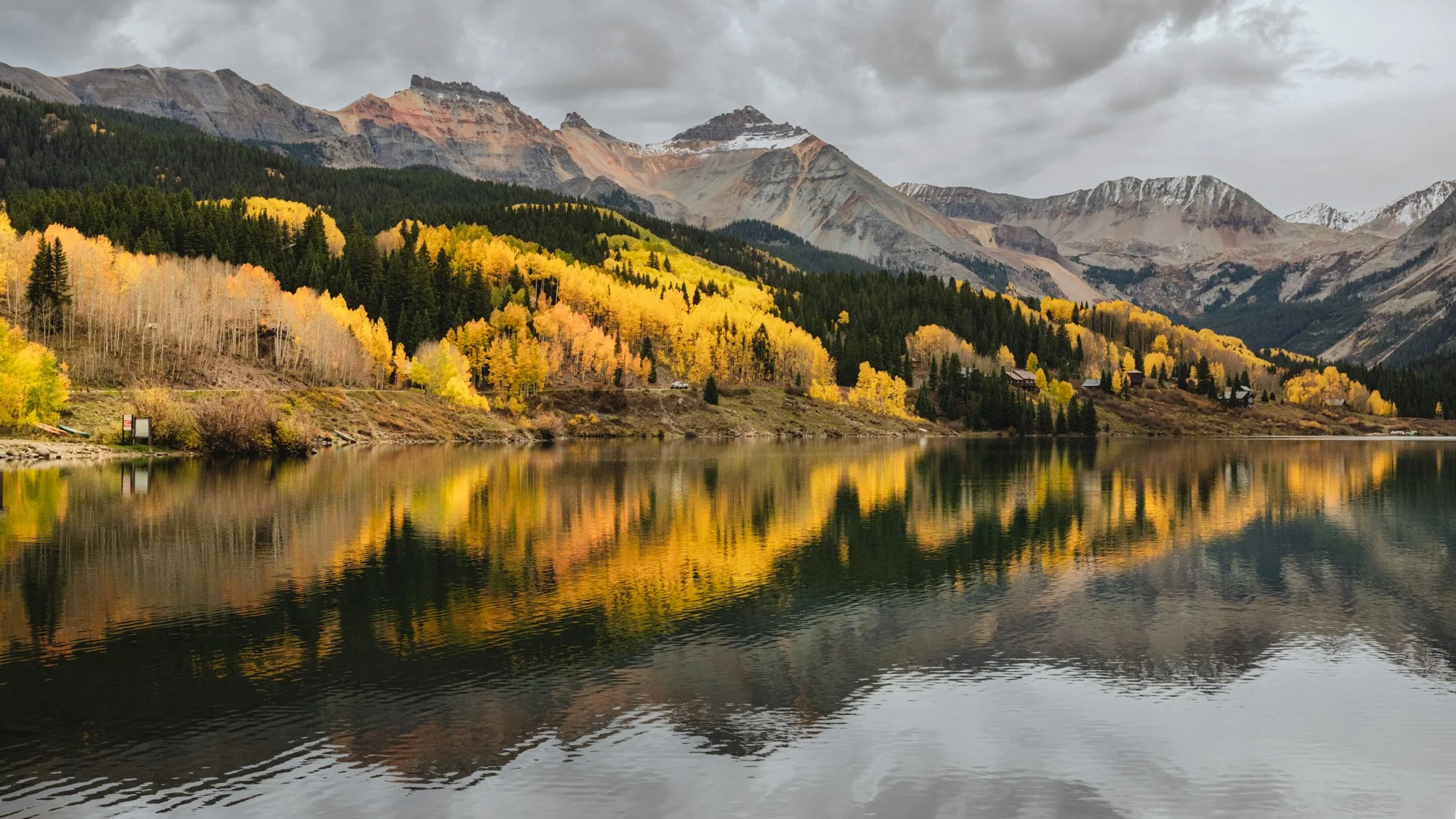 A mountain landscape with snow-capped peaks, colorful fall foliage of yellow, orange, and green trees along the shoreline of a calm lake, reflecting the scenery, under a cloudy sky.
