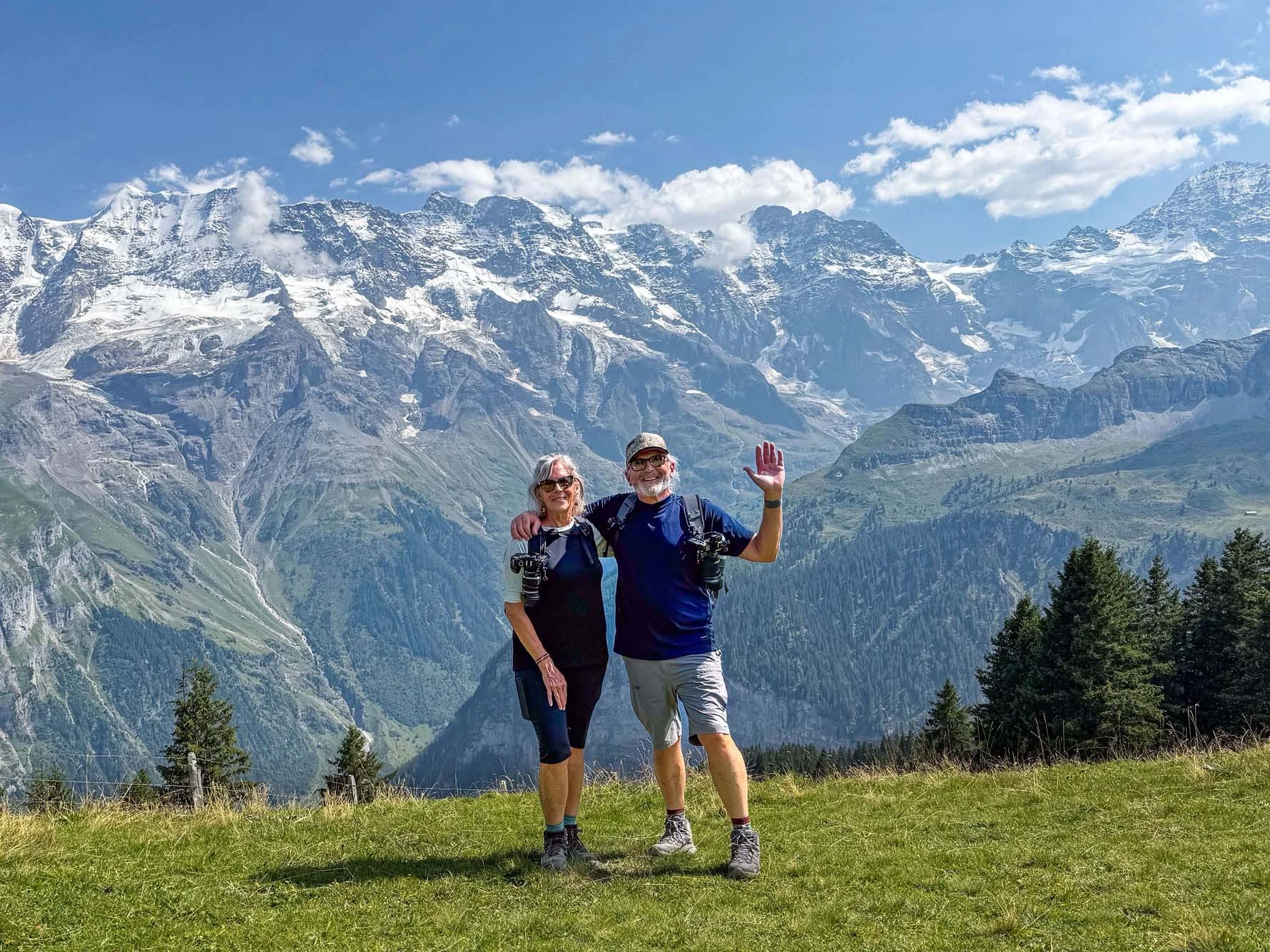 A smiling elderly couple standing on grass in front of snow-capped mountains, wearing hiking gear, with trees in the foreground and a clear blue sky.