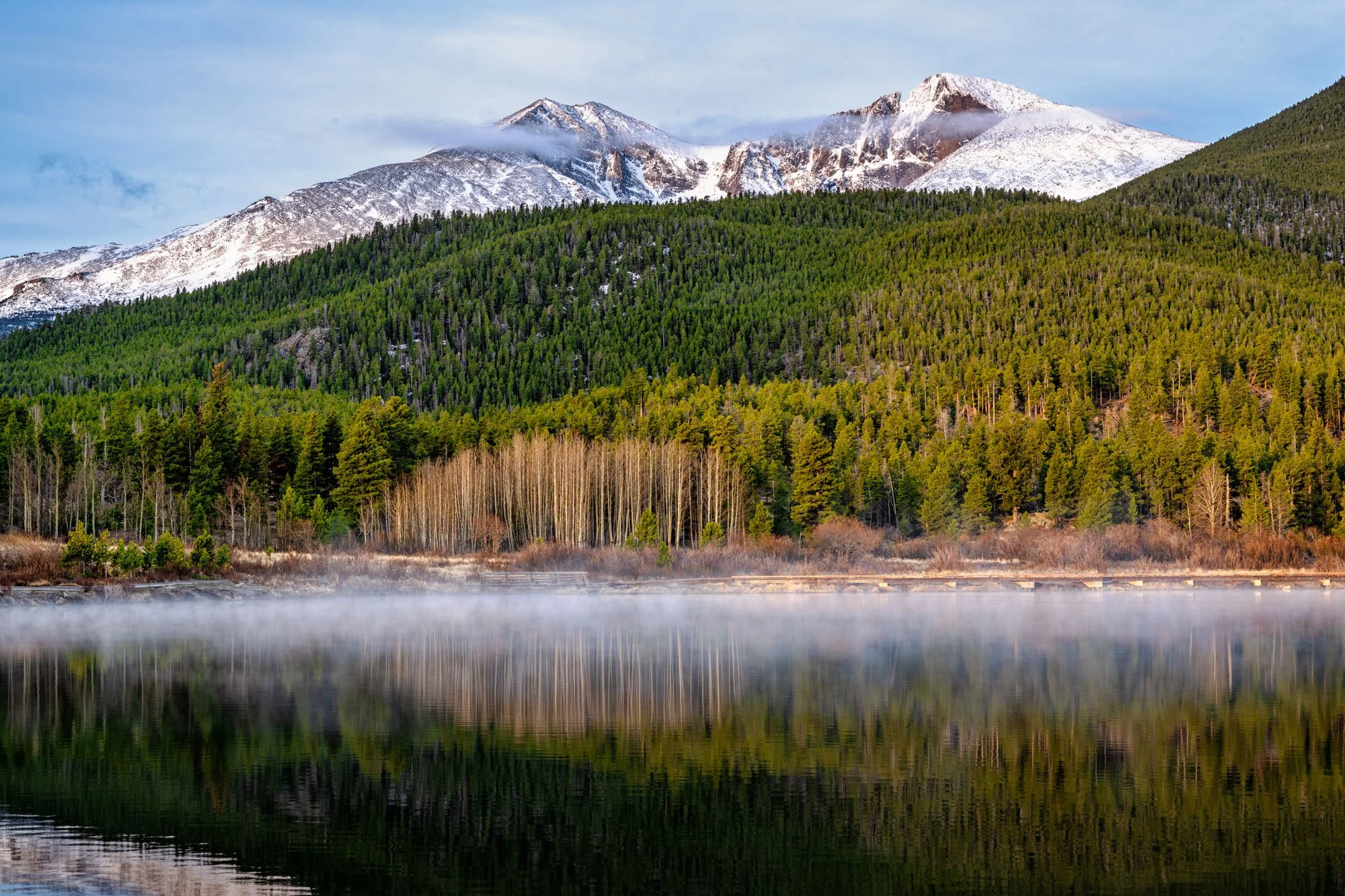 Snow-capped mountains in Colorado overlooking a dense green forest and a calm lake with mist on the surface.