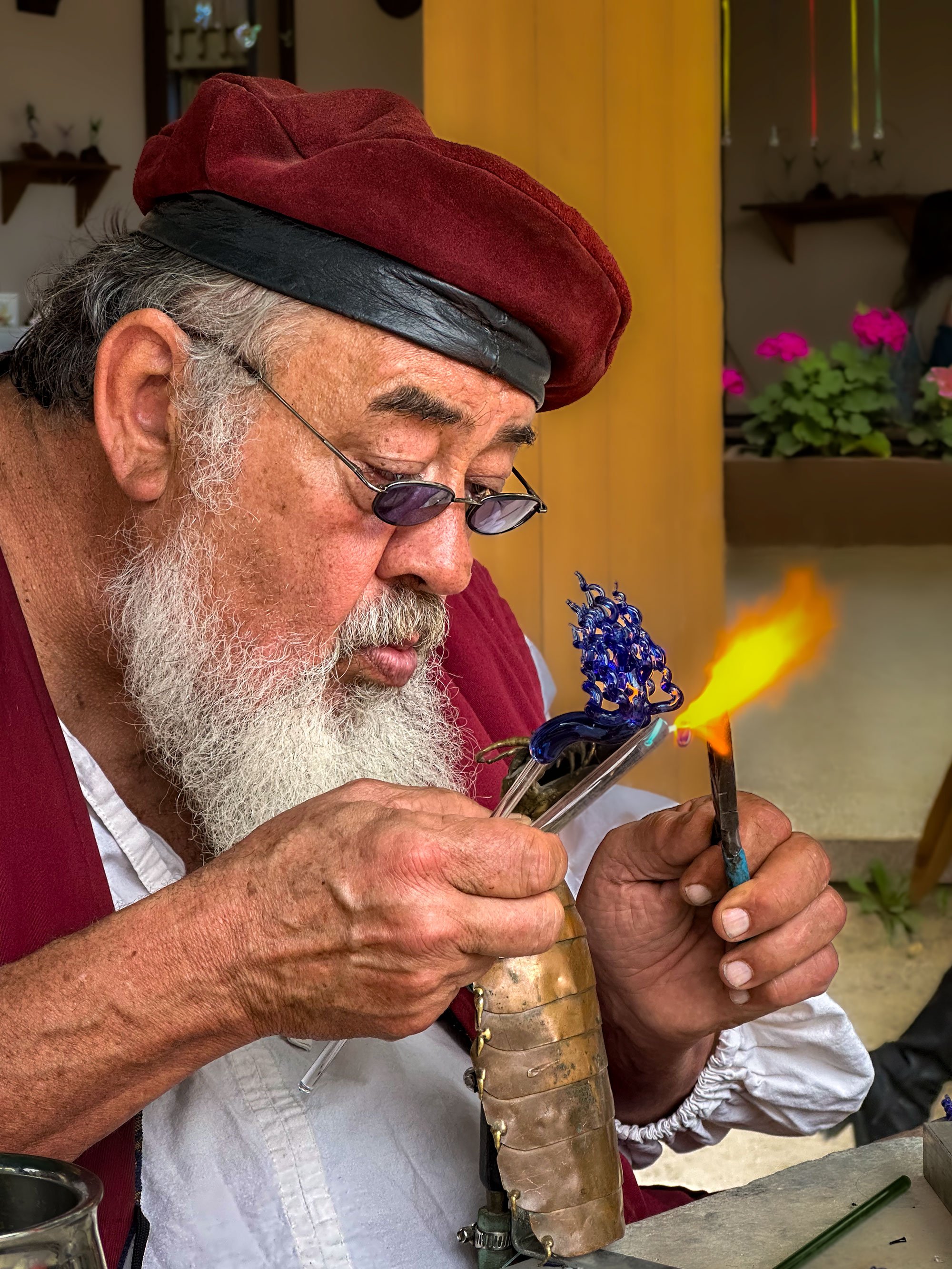 An elderly man with a white beard, wearing glasses and a red cap, is working with a blue glass sculpture and torch in his hand, during a glassblowing process in an indoor workshop.