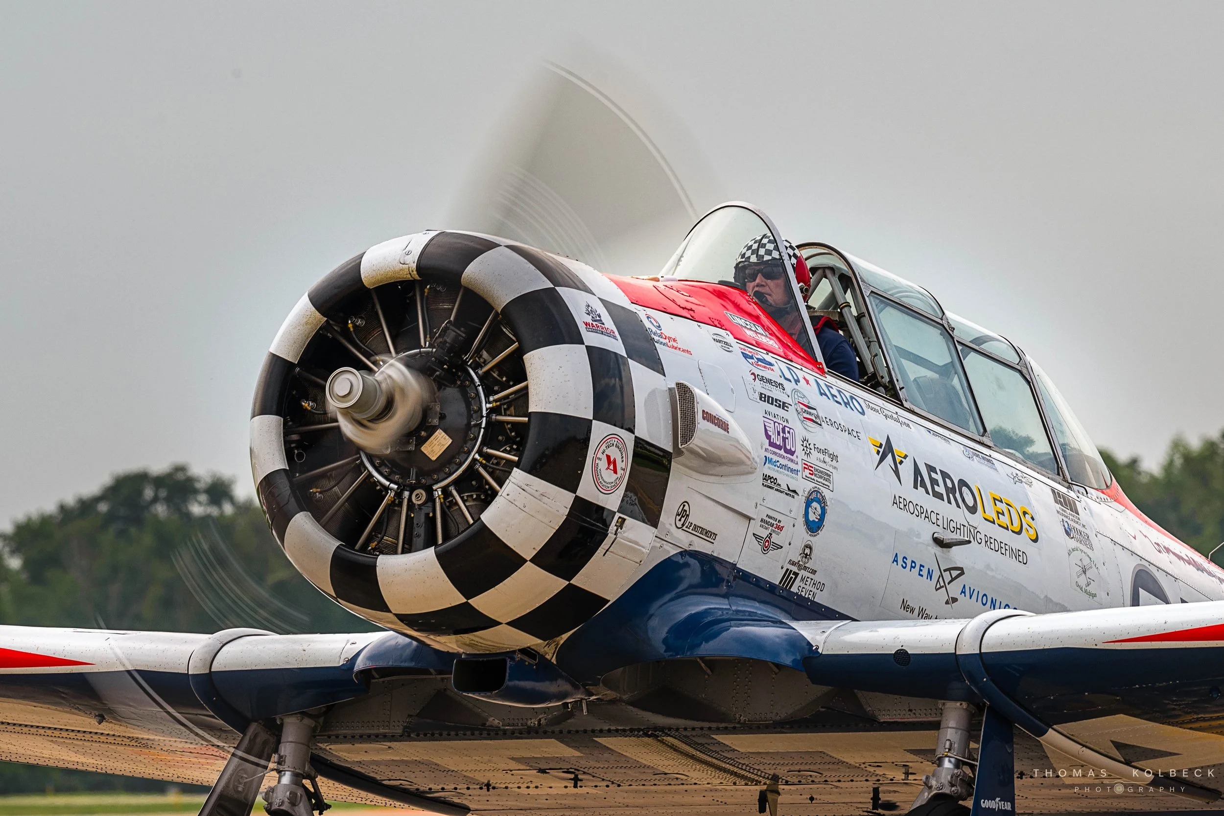 A vintage aircraft in motion, featuring a propeller with a black and white checkered pattern on the nose cone, and the cockpit with a pilot wearing a checkered helmet. The plane is decorated with various sponsorship logos.