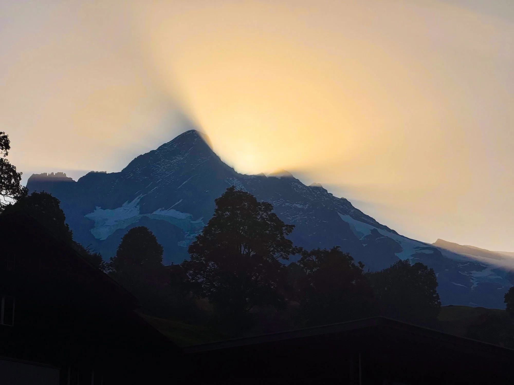 Sunset behind a mountain peak with clouds and trees in the foreground.