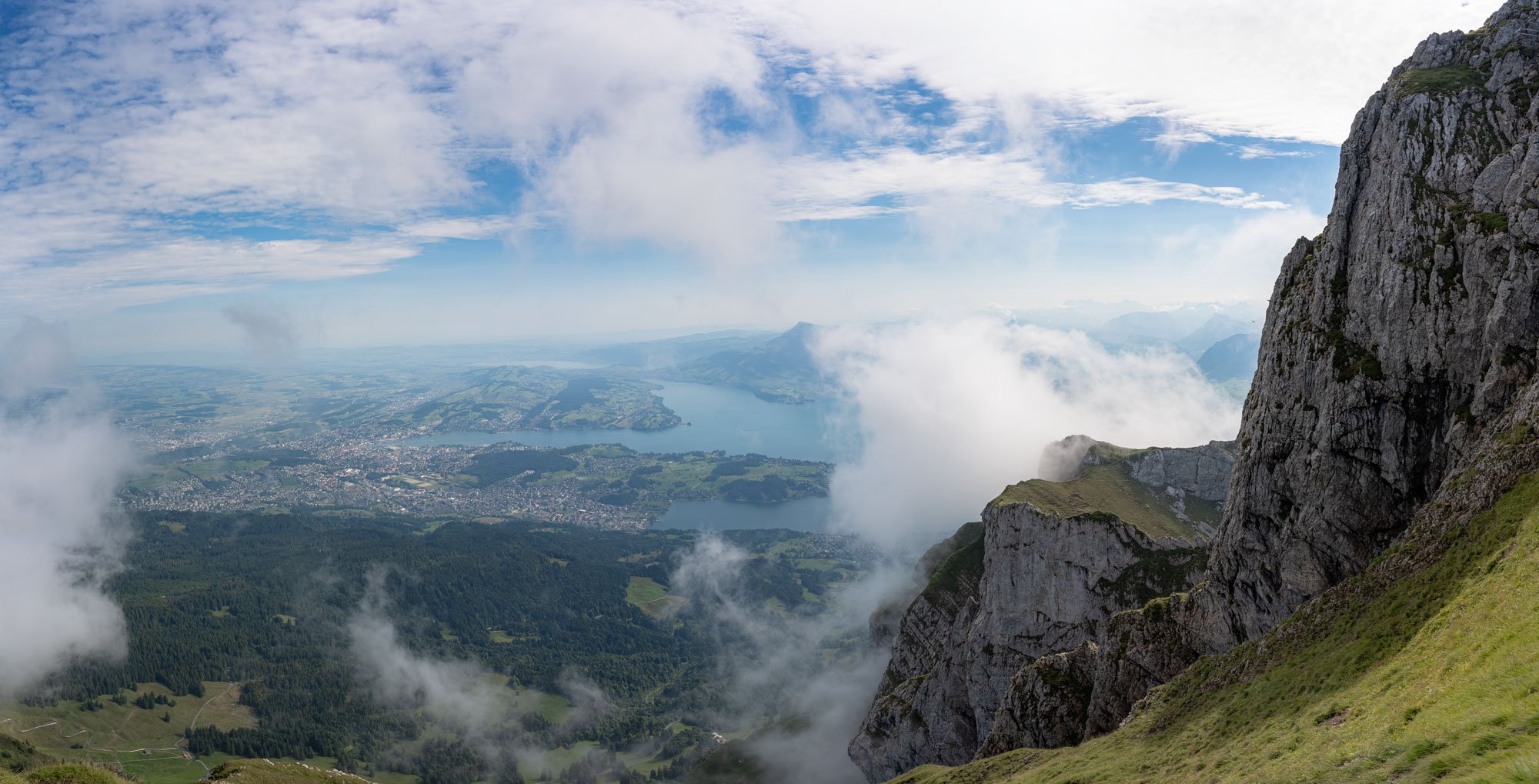 A scenic mountain view with a steep rocky cliff on the right, green grassy slopes, and clouds partially covering the landscape. Below is a large lake with surrounding land, and the sky is partly cloudy.