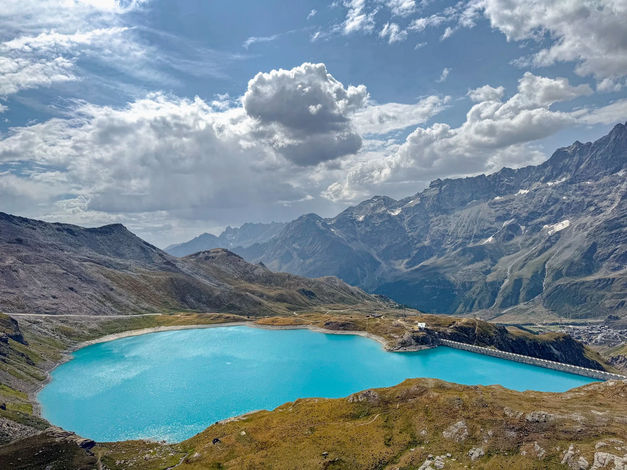 A scenic mountain landscape with a bright blue lake in the foreground, surrounded by rocky and grassy terrain, with rugged mountains in the background under a partly cloudy sky.
