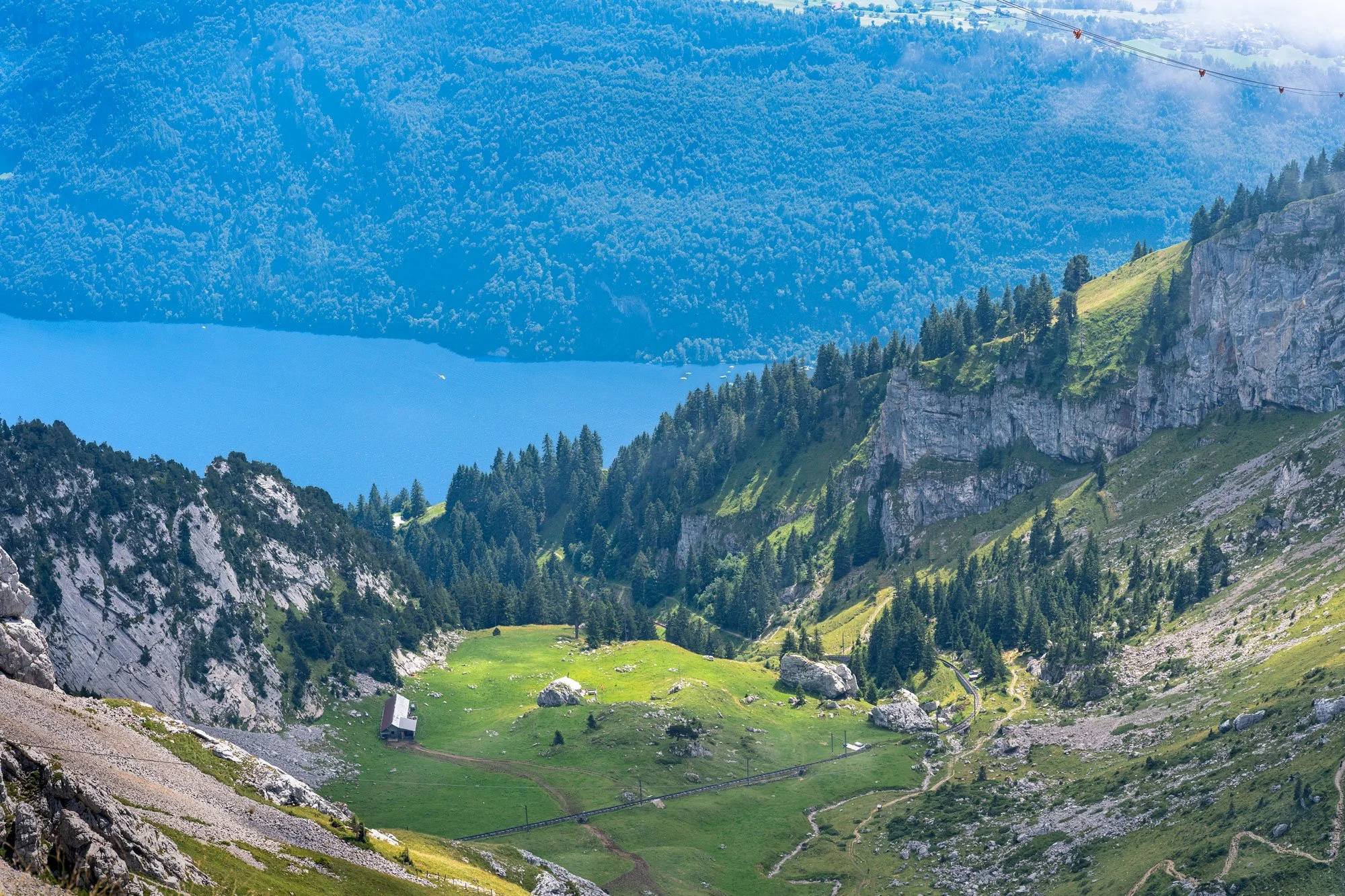 A scenic mountain landscape with green grassy fields, large rocks, dense forested areas, and a lake in the distance, under a partly cloudy sky.