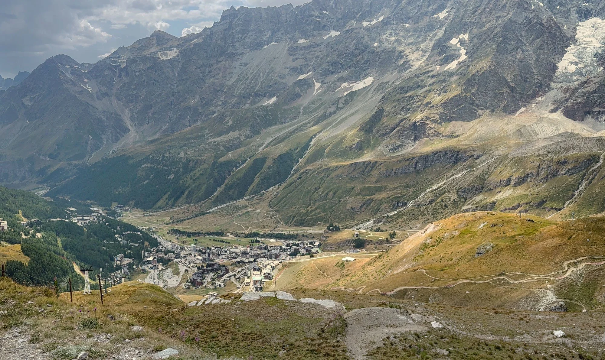 Mountain landscape with a small town at the valley, winding roads, and green and rocky slopes with patches of snow on the peaks in the background.