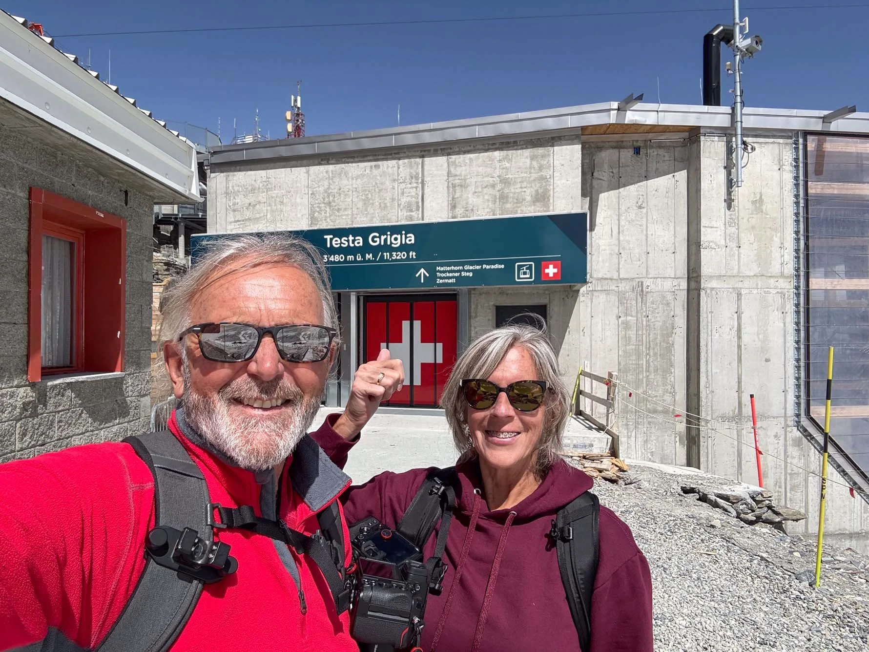 A smiling older man and woman in sunglasses and outdoor gear taking a selfie in front of a sign for Testa Grigia at a high altitude in the Alps, with a concrete building and alpine landscape in the background.