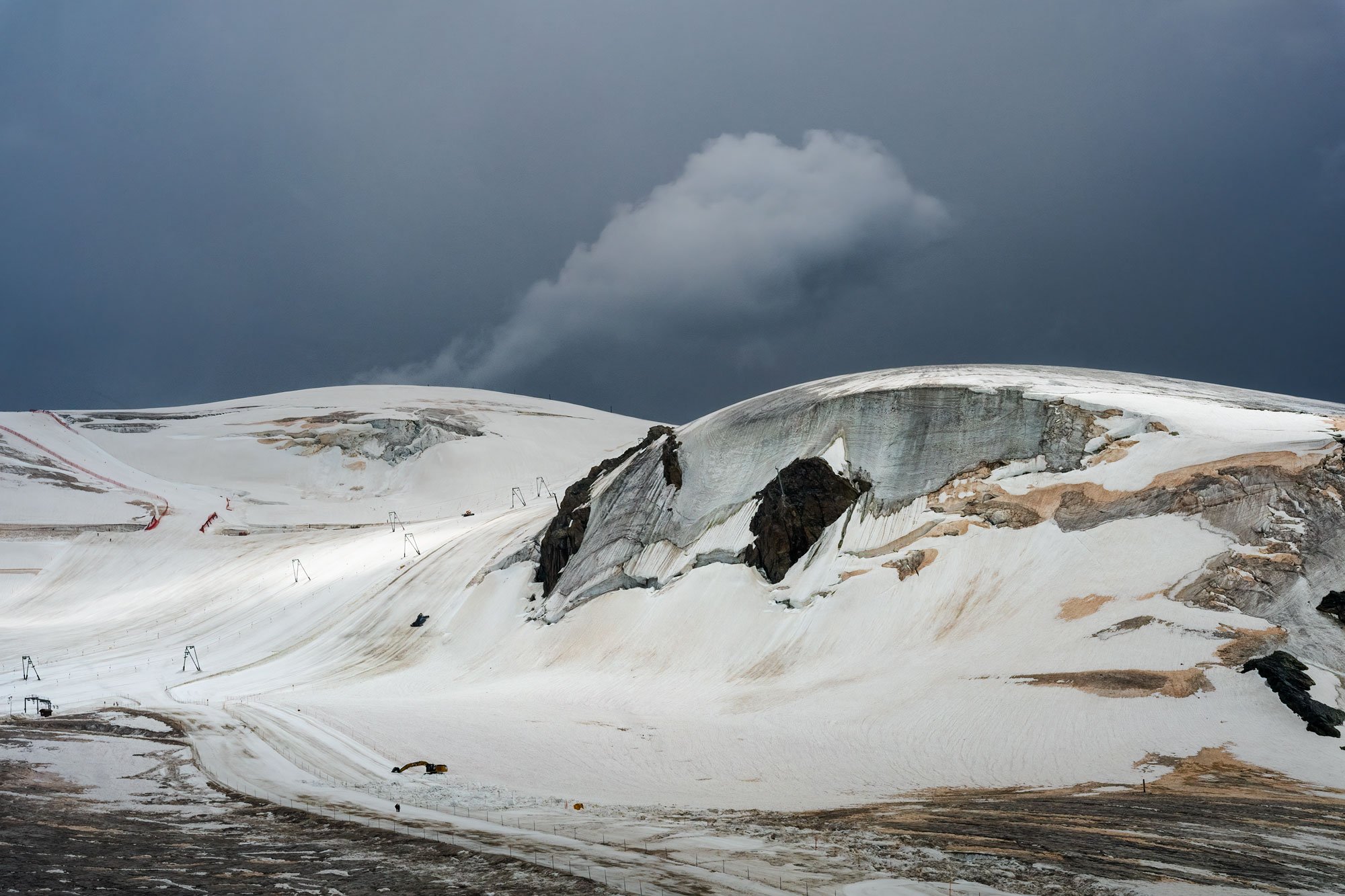 Snow-covered mountain landscape with dark stormy clouds in the sky, a glacier, and some construction equipment.