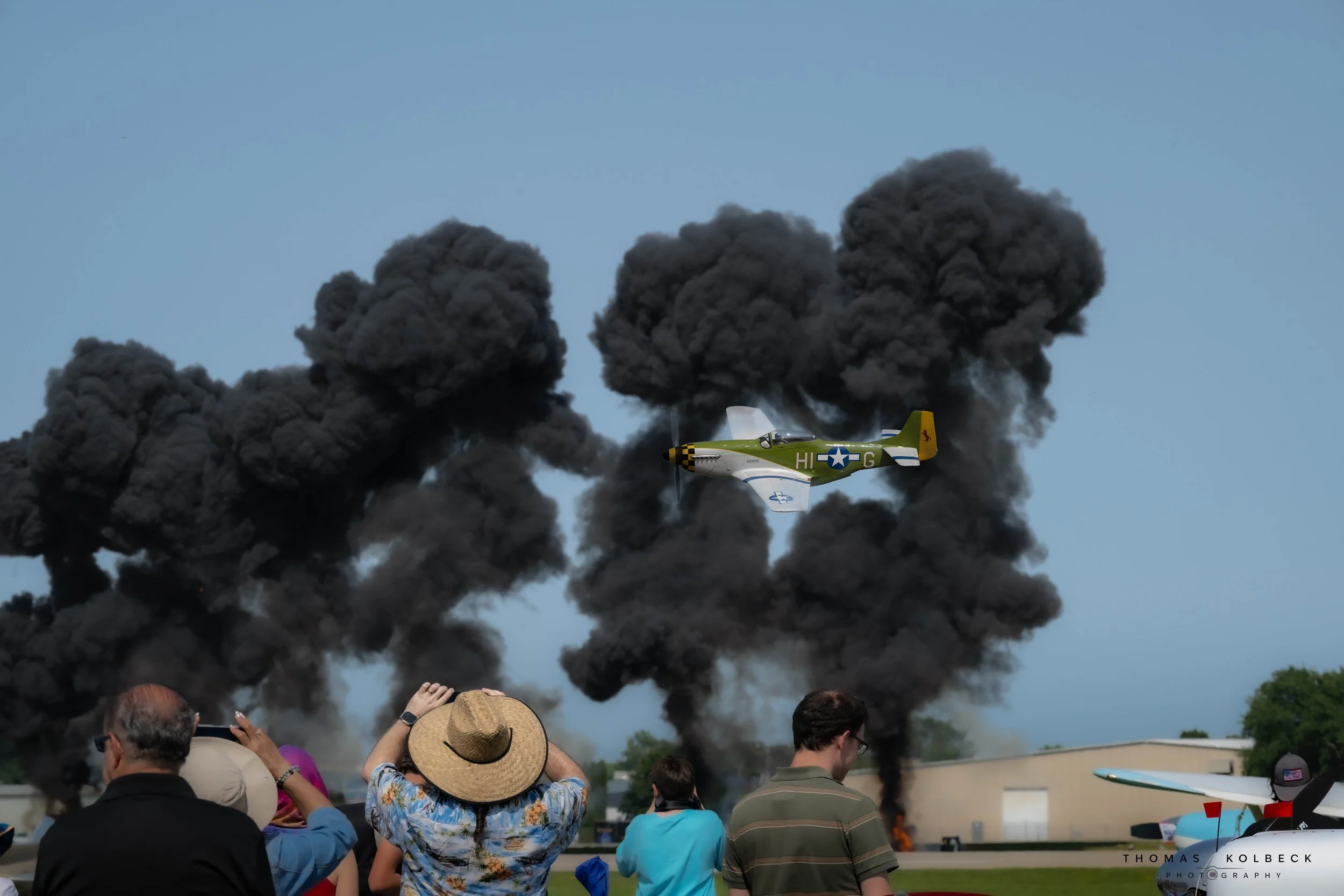 People watching a vintage airplane flying low with black smoke trails behind it during an air show, with large plumes of black smoke rising from fires on the ground in the background.