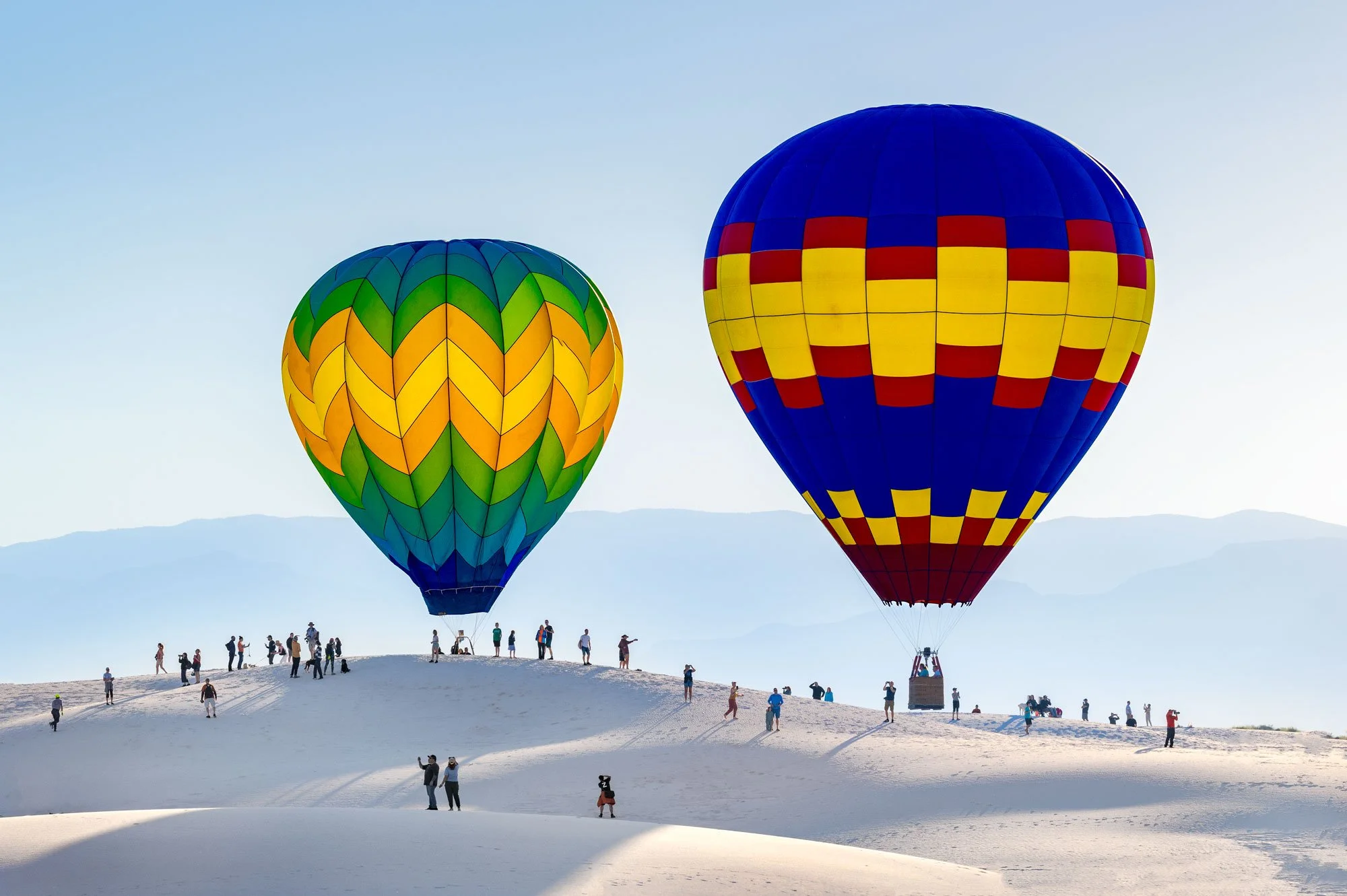 Two colorful hot air balloons flying over a white landscape with people standing and walking around