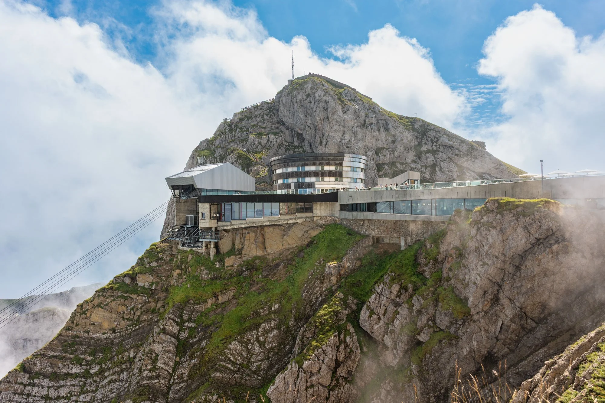 Mountaintop building with modern architecture and cable car cables, overlooking clouds and rocky cliffs.