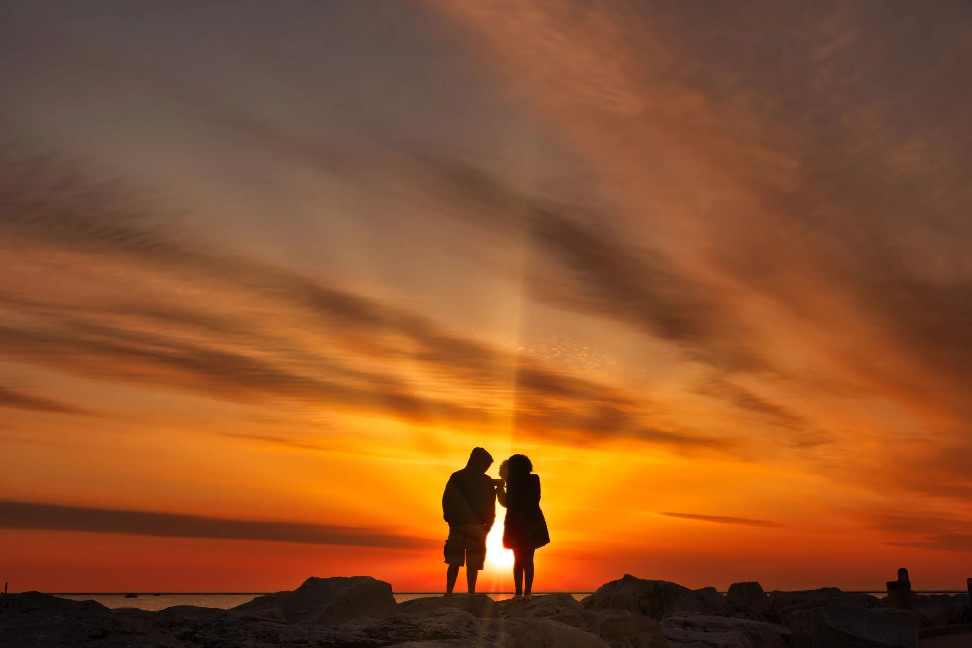 Silhouette of a couple standing on rocks at sunset with orange sky and clouds.