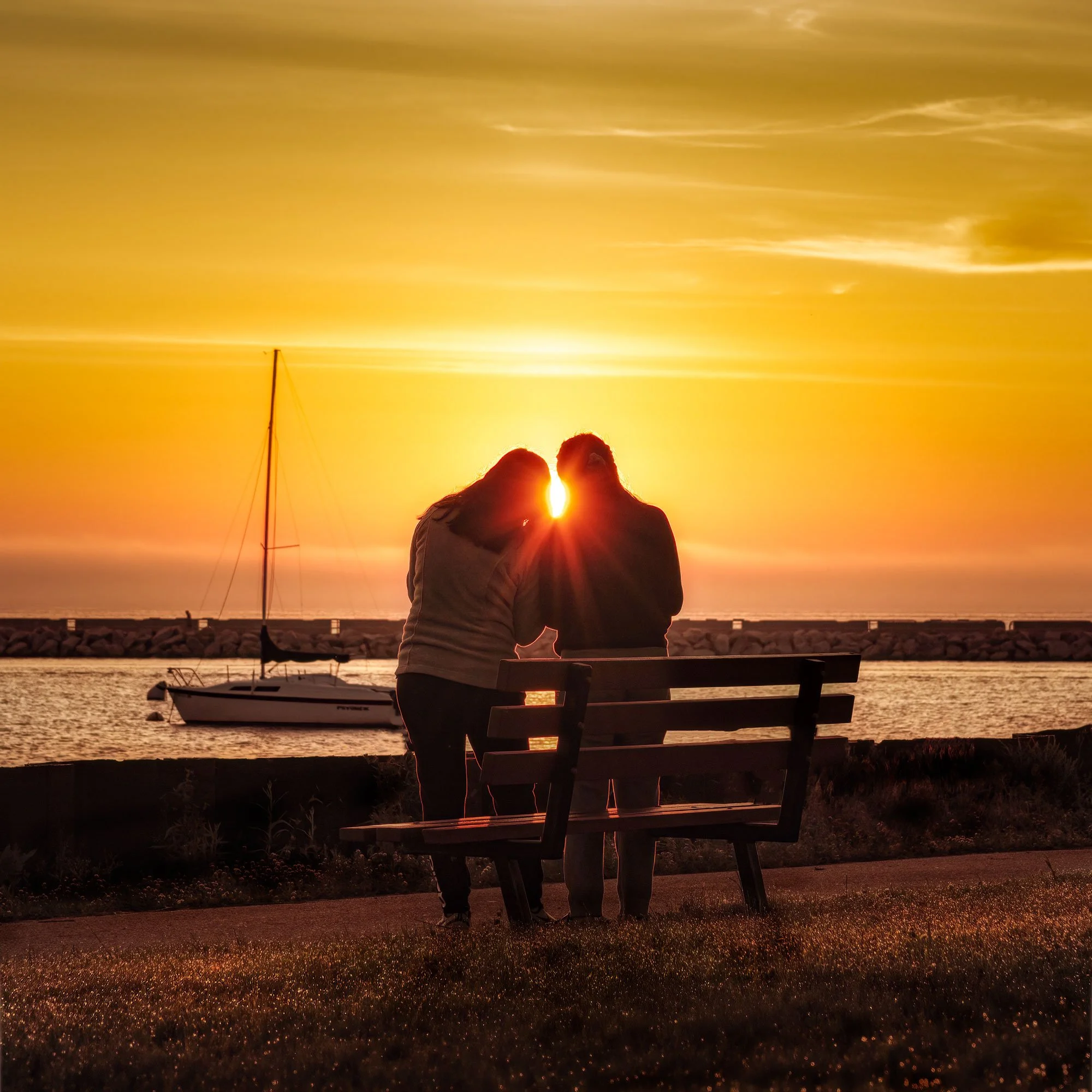 Couple by a park bench at sunset by the water, with sailboat and sailboat mast in the background.