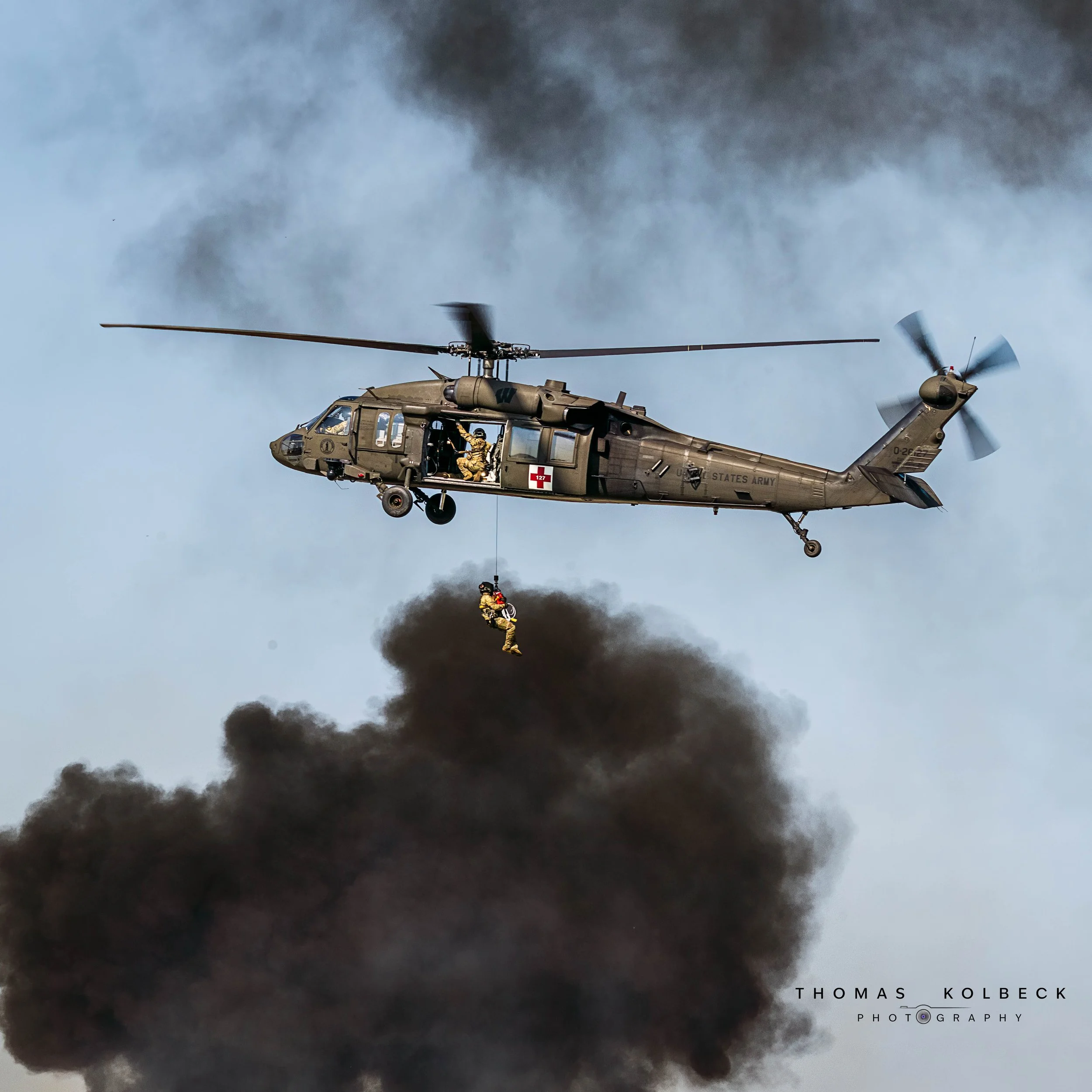 Military helicopter with soldiers, one hanging underneath by a rope, amid smoke clouds.