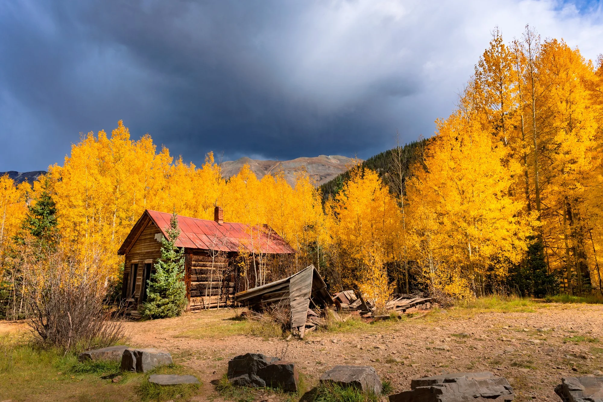 An old wooden house with a red metal roof, surrounded by yellow autumn trees and rocky ground, under a cloudy blue-sky mountain backdrop.