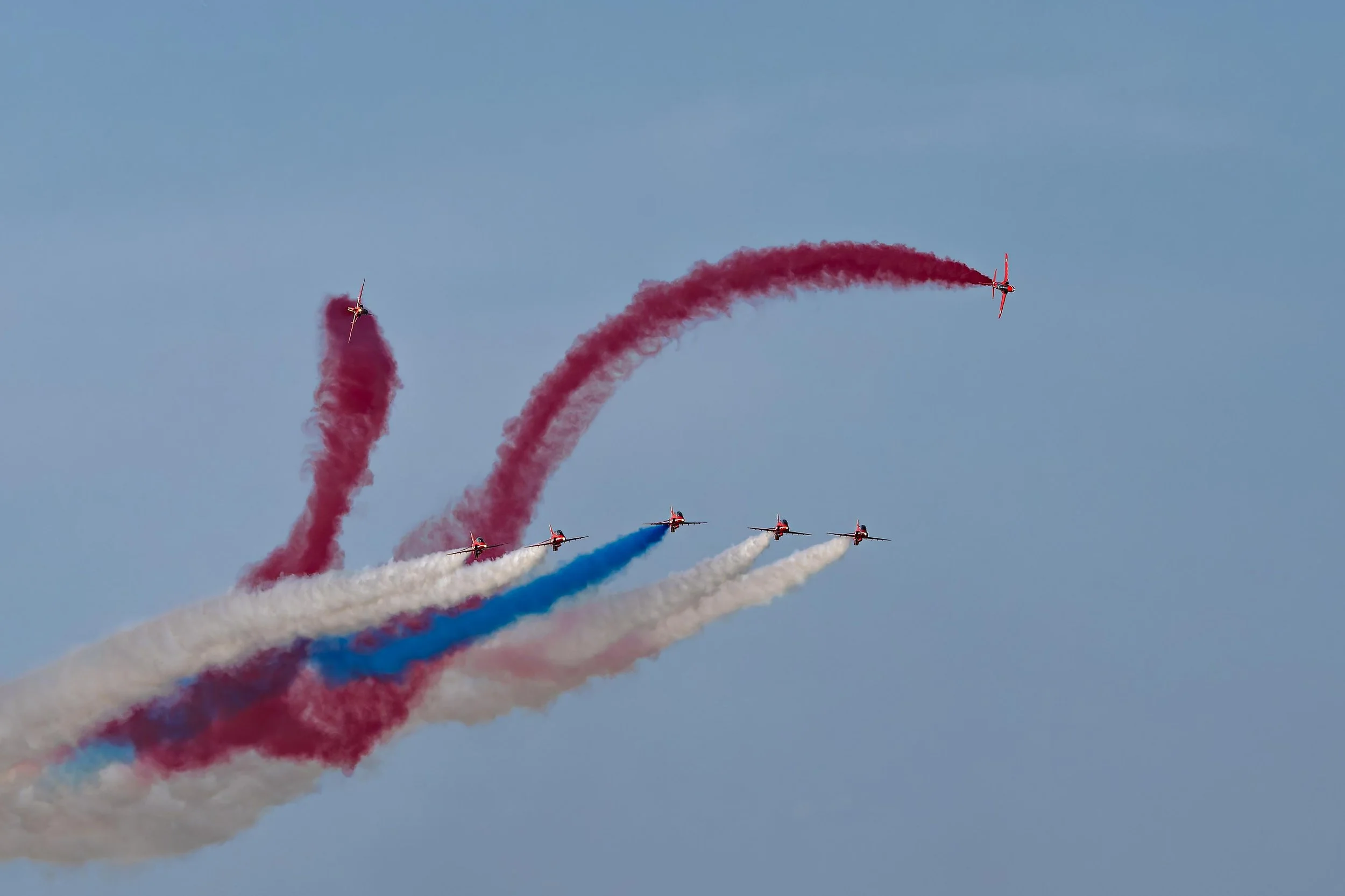 Six aircraft performing an aerobatic flight in formation with trails of colored smoke—red, white, and blue—in the sky.