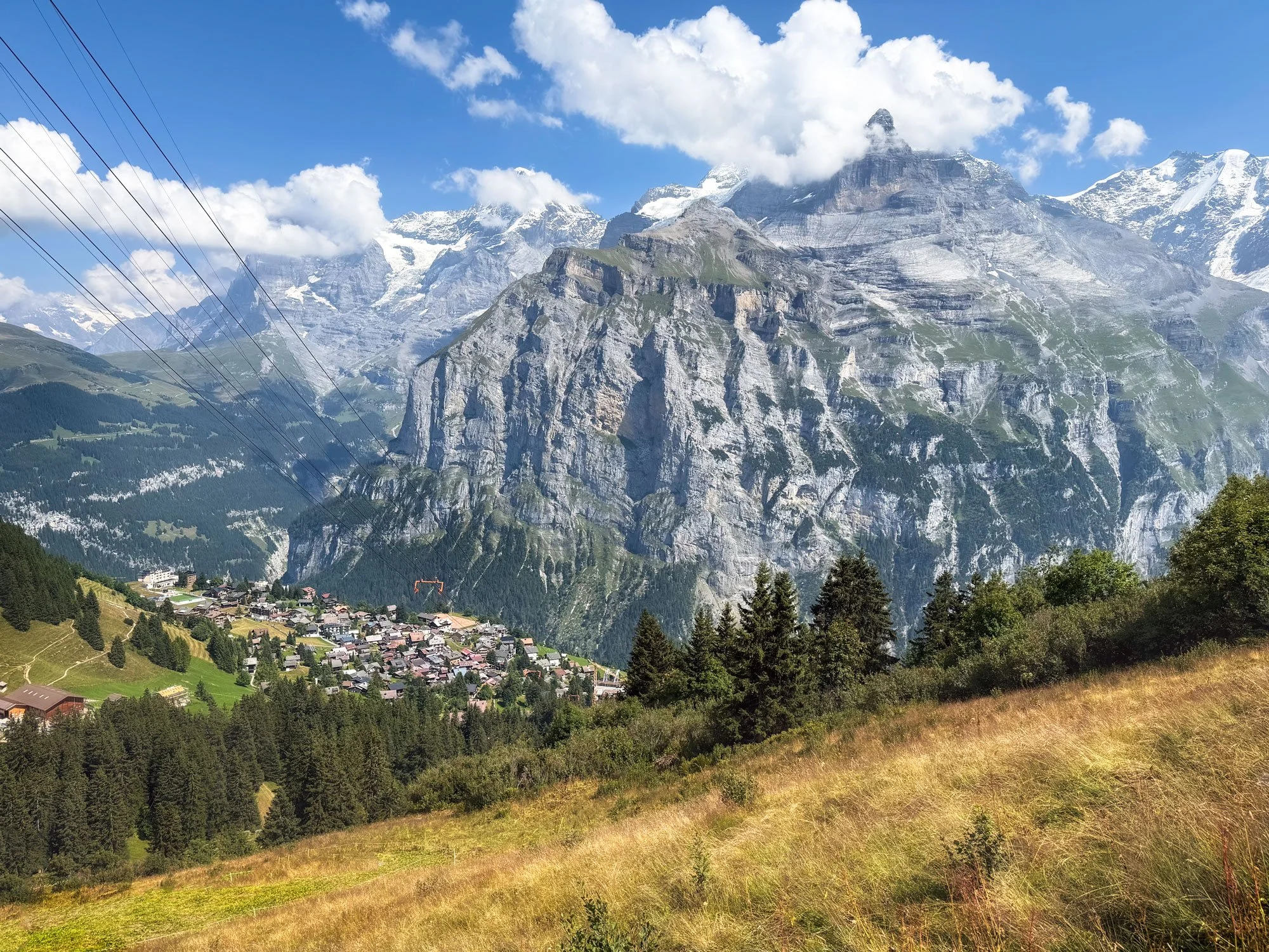 Mountain landscape with a village at the base, featuring green trees, grassy fields, and towering snow-capped peaks under a partly cloudy sky.