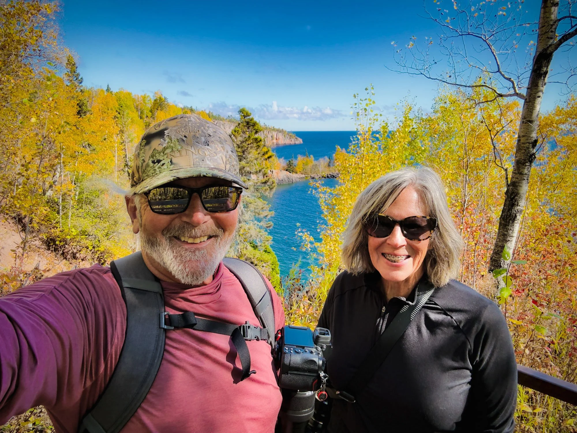 Smiling man and woman taking a selfie outdoors during fall, surrounded by yellow and orange trees with a lake and coast in the background.