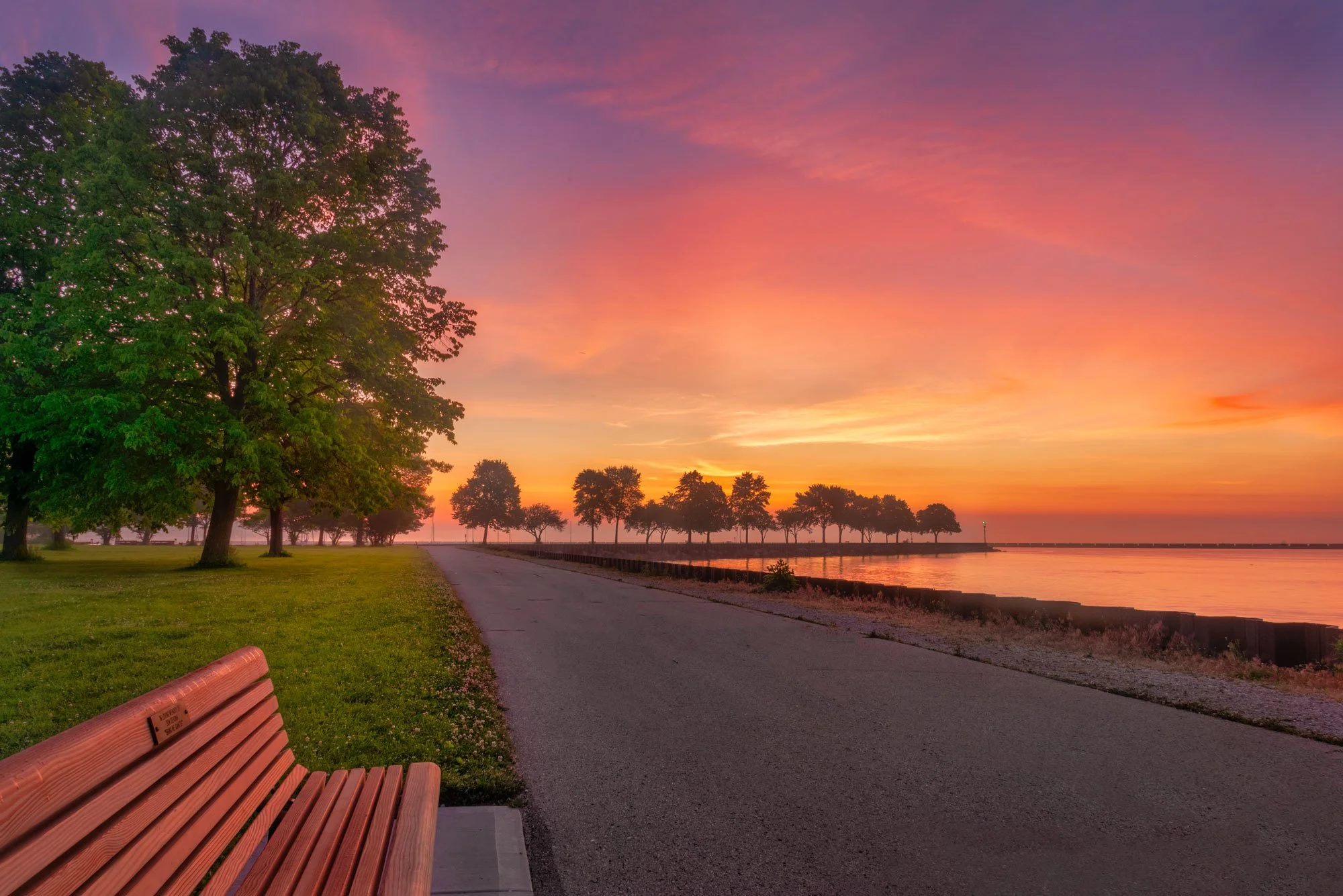 A park path at sunset with a bench on the left, green trees on the left, and a calm body of water on the right, all under a colorful sky with pink, purple, and orange hues.