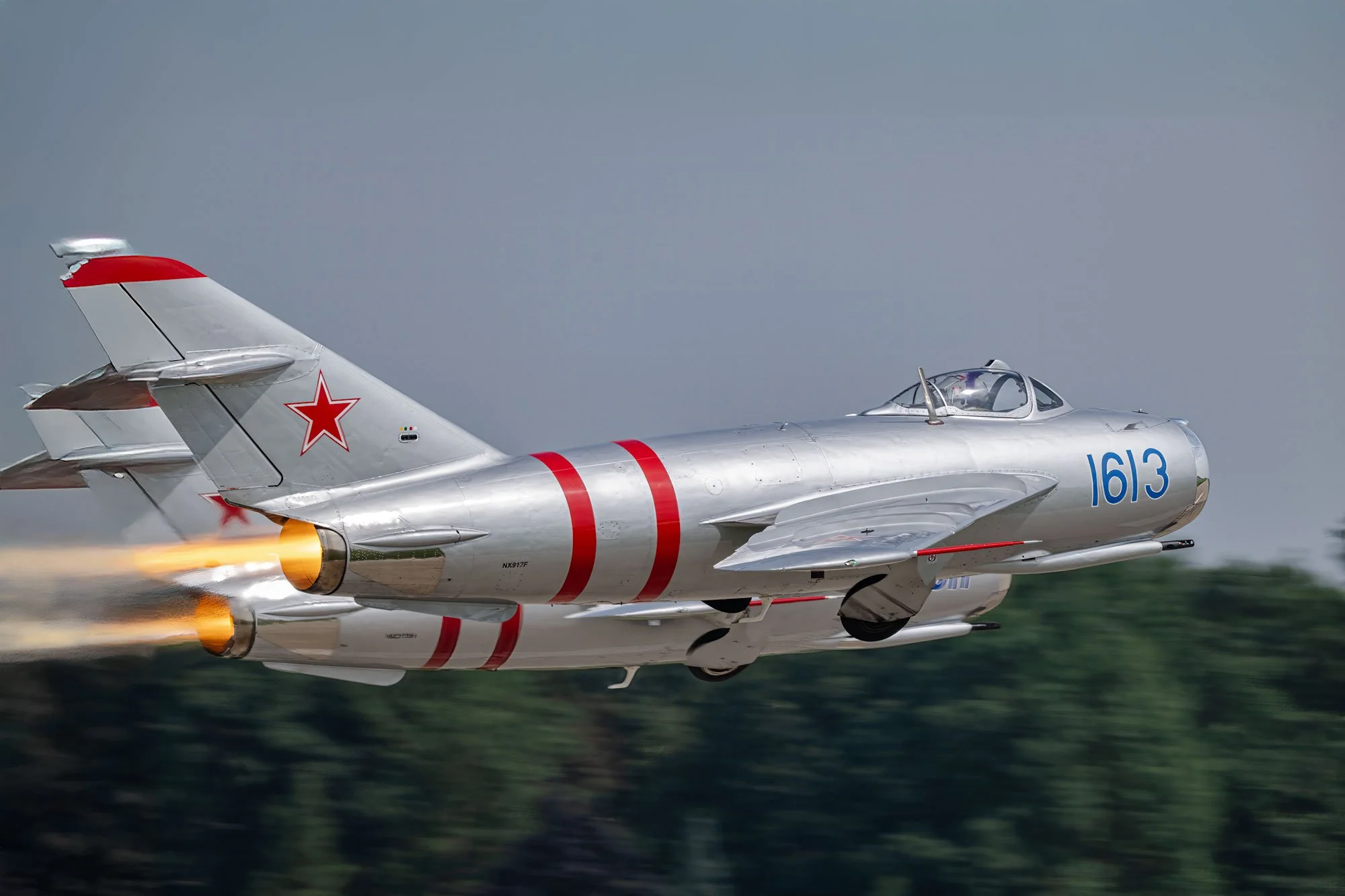 A silver military jet with red stars on the tail and blue number 1613 on the nose, flying at high speed with flames coming out of its engines.