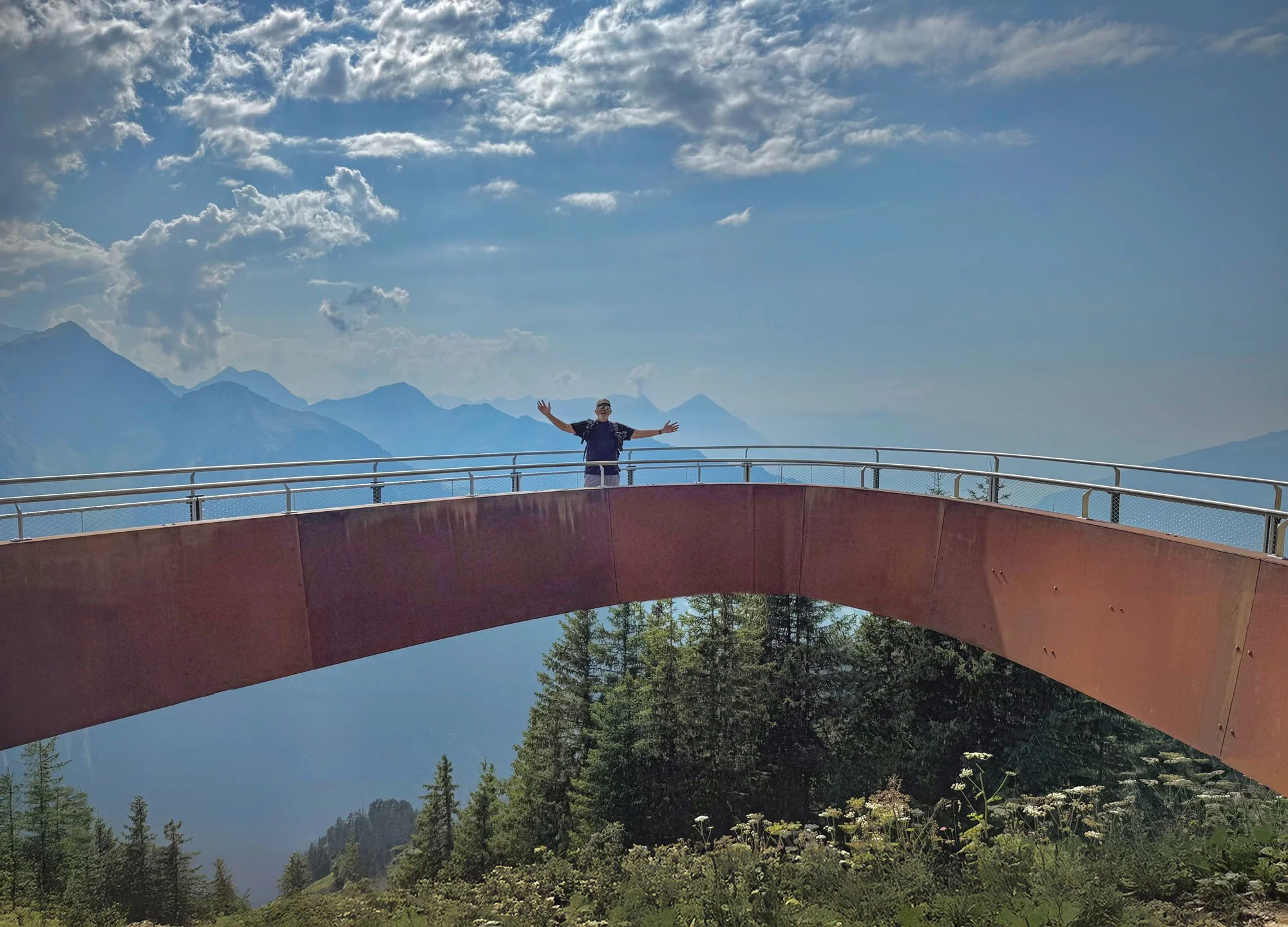 A person standing on a curved mountain lookout bridge with arms outstretched, surrounded by a forest and mountain range under a partly cloudy sky.
