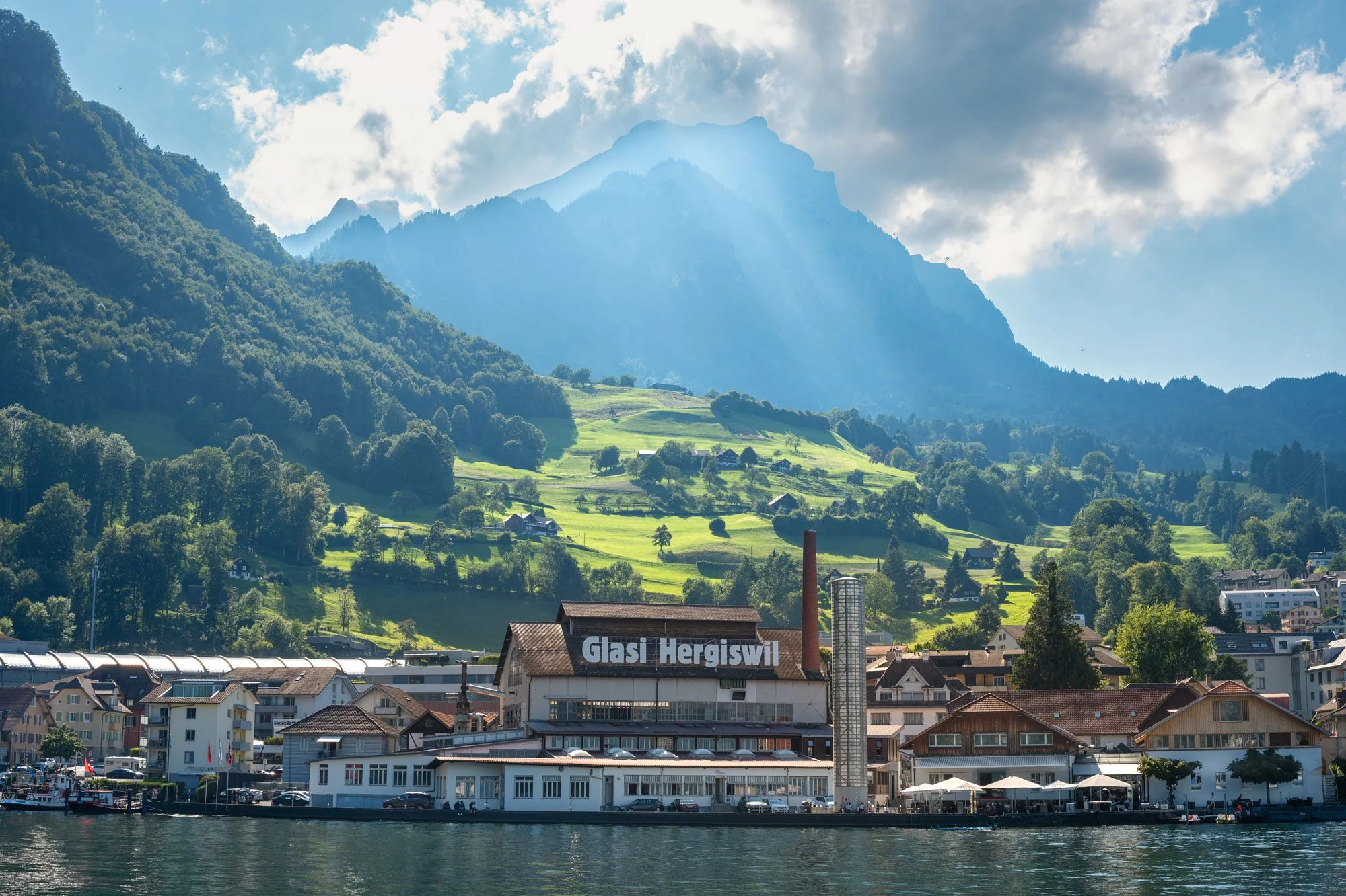 A scenic lakeside view with a building labeled 'Glasi Hergiswil' in the foreground, surrounded by houses, with lush green hills and mountains in the background under a partly cloudy sky.
