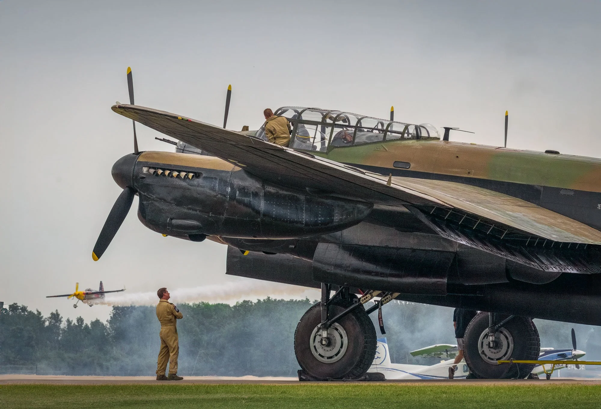 A military aircraft on the ground with ground personnel nearby. In the background, a smaller plane is flying, releasing white smoke.