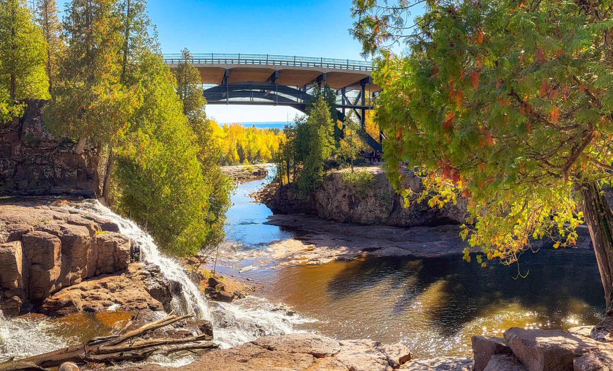 A scenic view of a river surrounded by lush trees with fall foliage, a small waterfall on the left, and a bridge overhead in a forested area on a clear, sunny day.