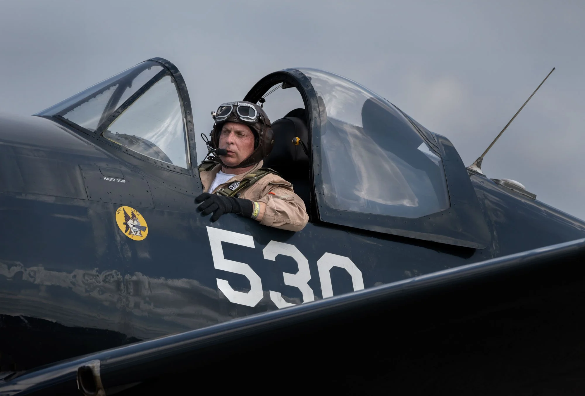 A male pilot sitting in the cockpit of a black fighter jet, wearing a helmet with goggles and a beige flight suit, with a cloudy sky in the background.