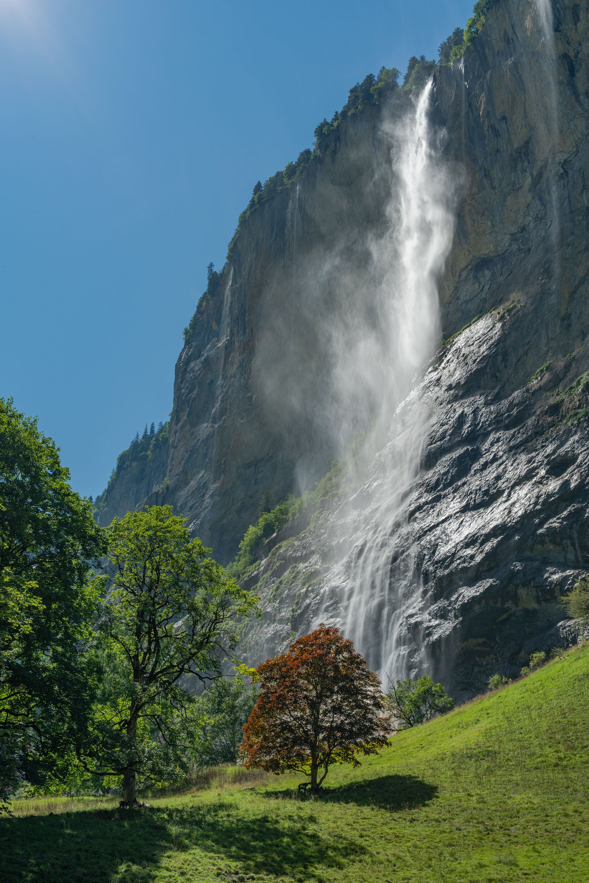 Tall waterfall flowing down a rocky mountain, surrounded by green trees and a grassy hill under a clear blue sky.
