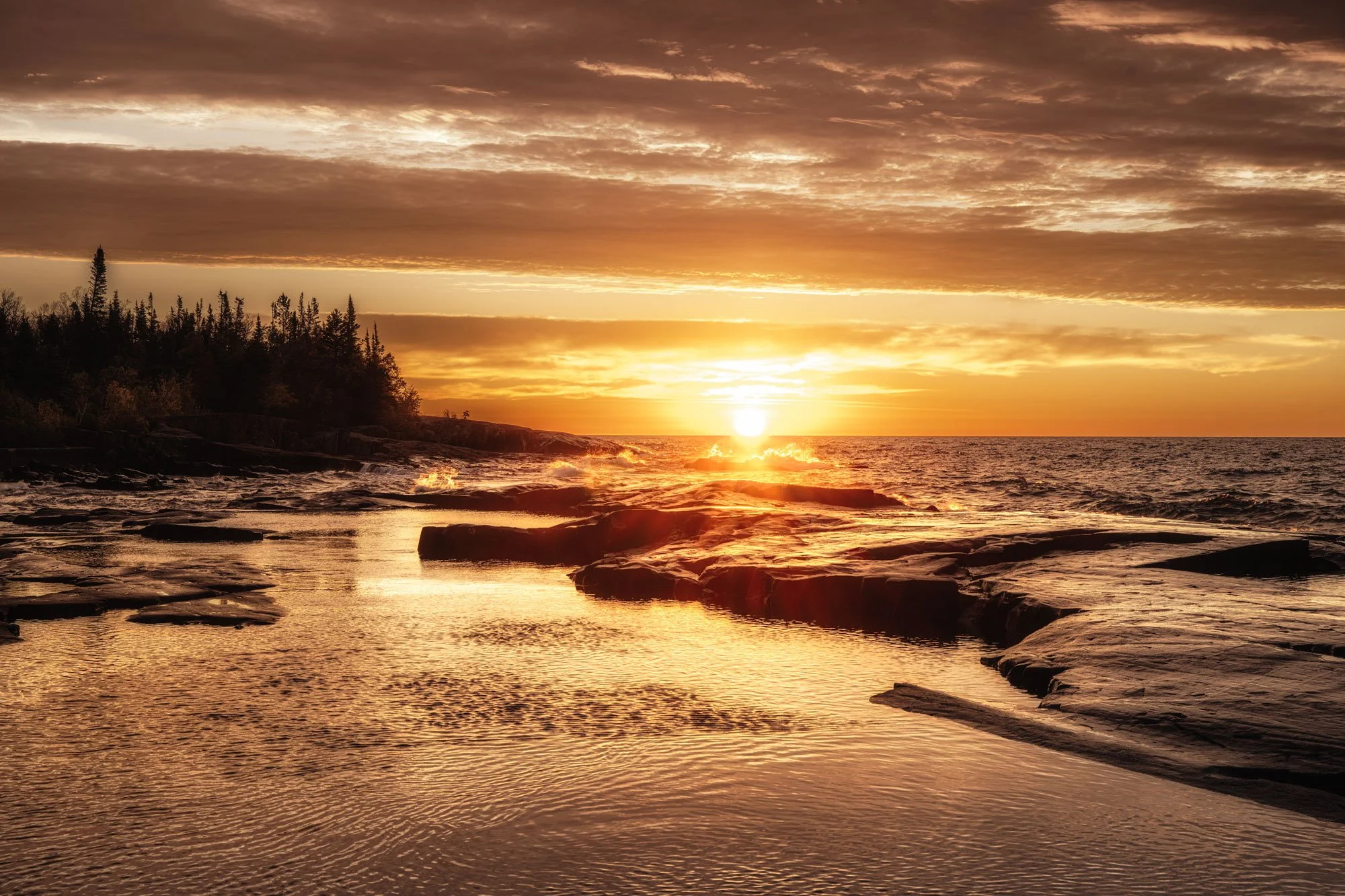 A sunset over a rocky shoreline with water reflecting the orange sky and clouds, and a line of trees in the distance.