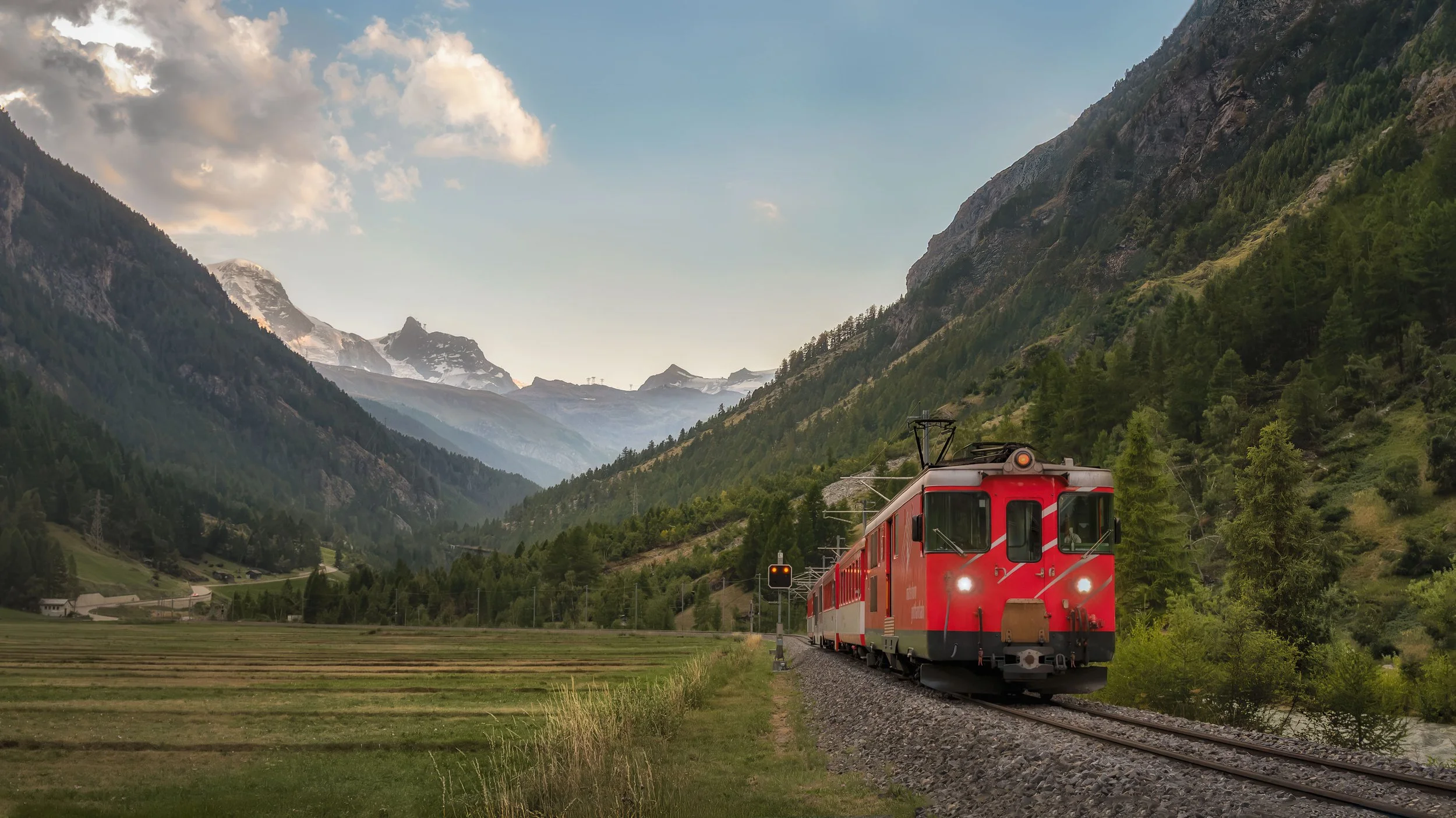 A red train moving through a scenic mountainous landscape with green hills, tall mountains, and trees, under a partly cloudy sky.