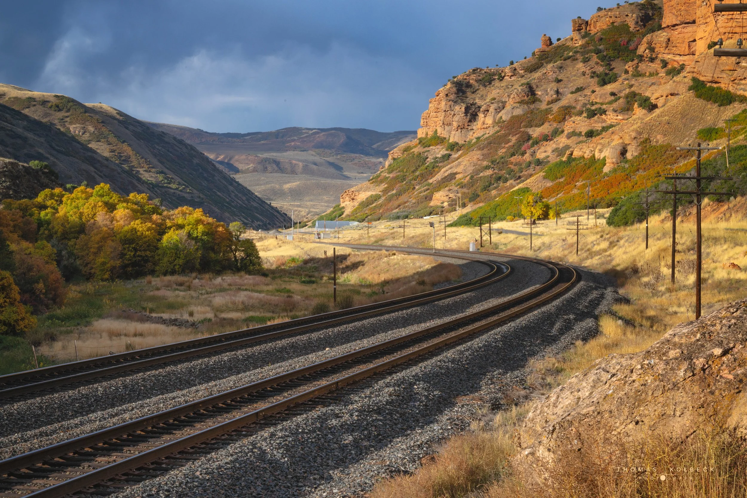 A scenic view of a train track curving through a desert valley with colorful autumn trees on the left and orange rocky hills on the right, under a partly cloudy sky.
