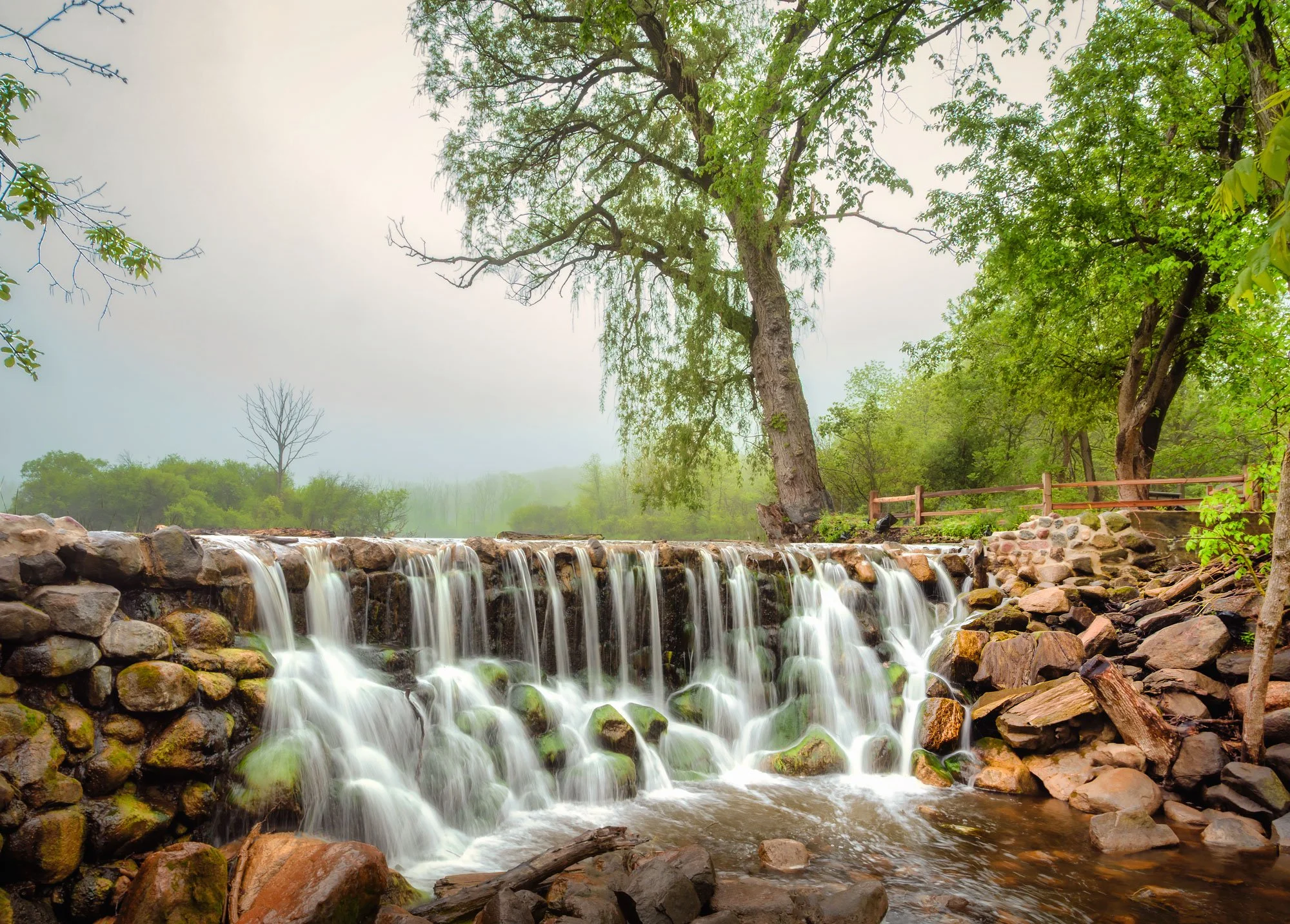 A small waterfall flowing over a rocky stone dam in a lush green forested area with trees and a wooden fence in the background.