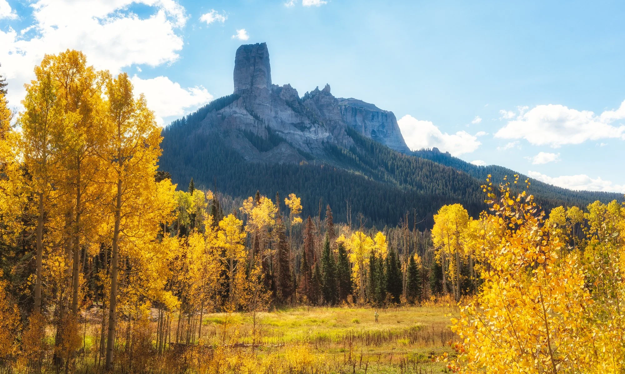 A landscape of a mountain with a tall rock formation in the background, surrounded by pine trees, with vibrant yellow and green fall foliage in the foreground, under a partly cloudy sky.