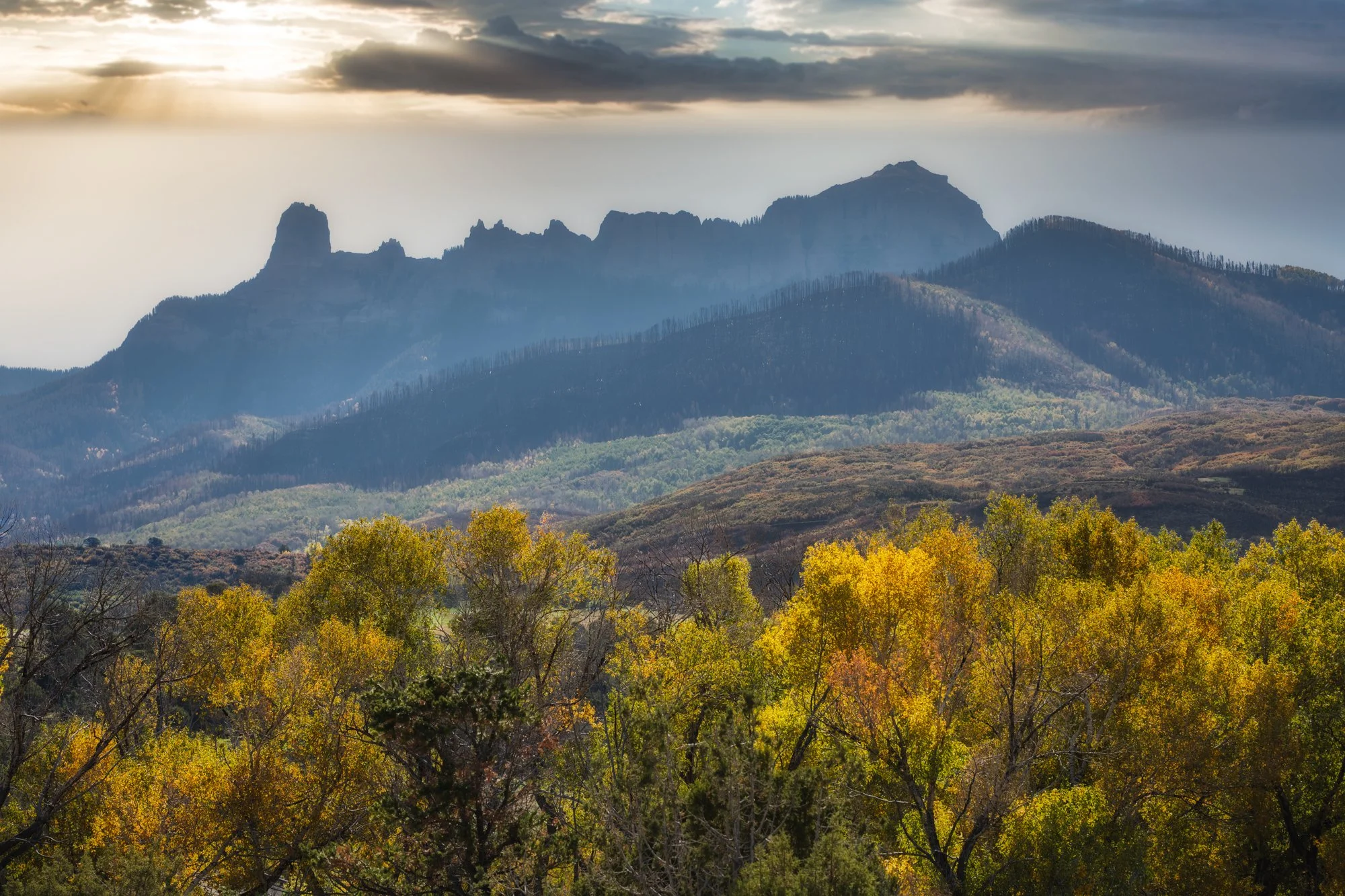 A landscape with mountains, some of which are jagged and others rounded, covered in forest with trees in fall colors of yellow, orange, and red, beneath a cloudy sky with sunlight breaking through.