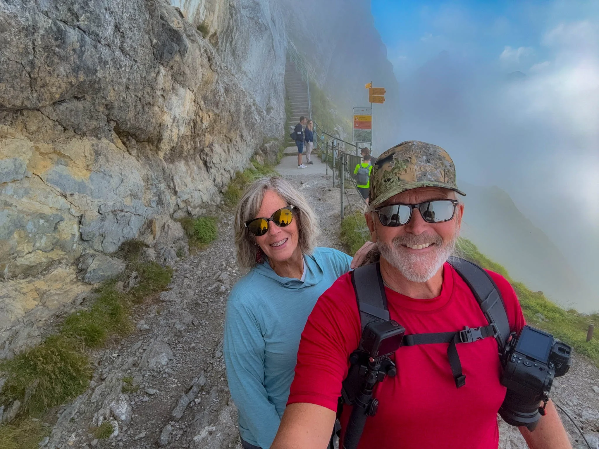 Smiling couple taking a selfie on a mountain trail with fog and mountains in the background.