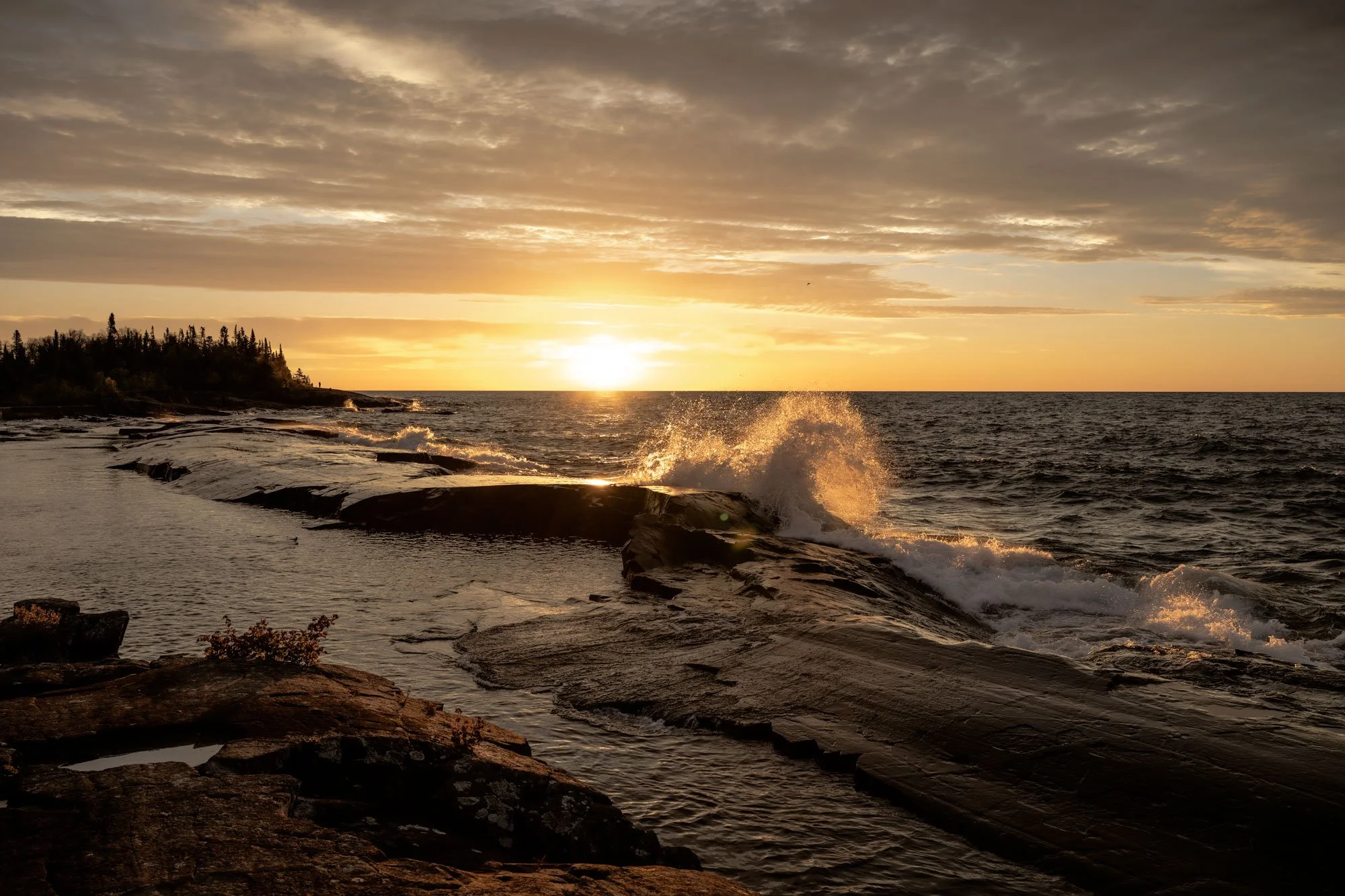 Sunset over a rocky shoreline with waves crashing, some trees on the distant left, and a cloudy sky.