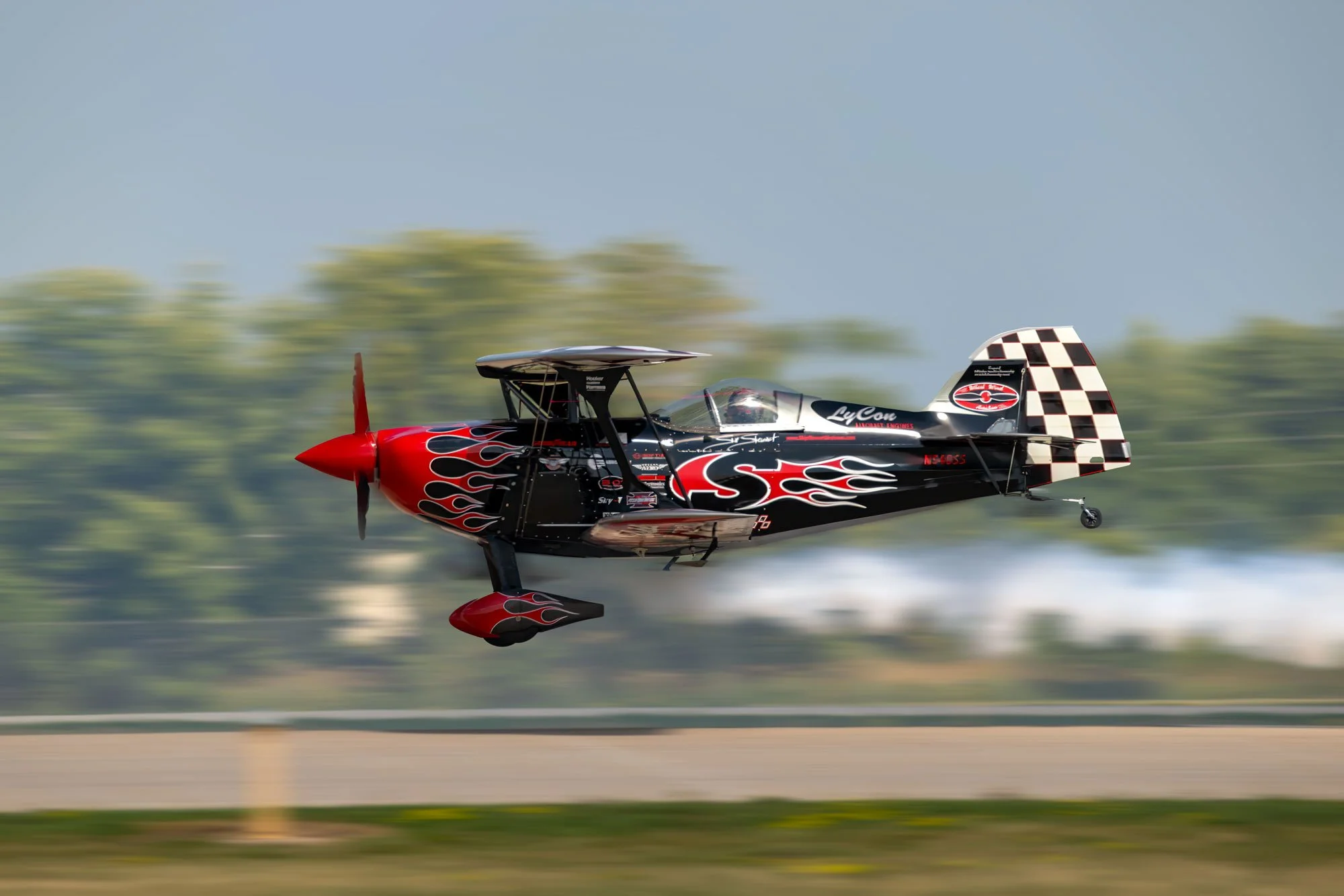 A black and red race plane with flame decals, flying low over a runway with blurred trees in the background.